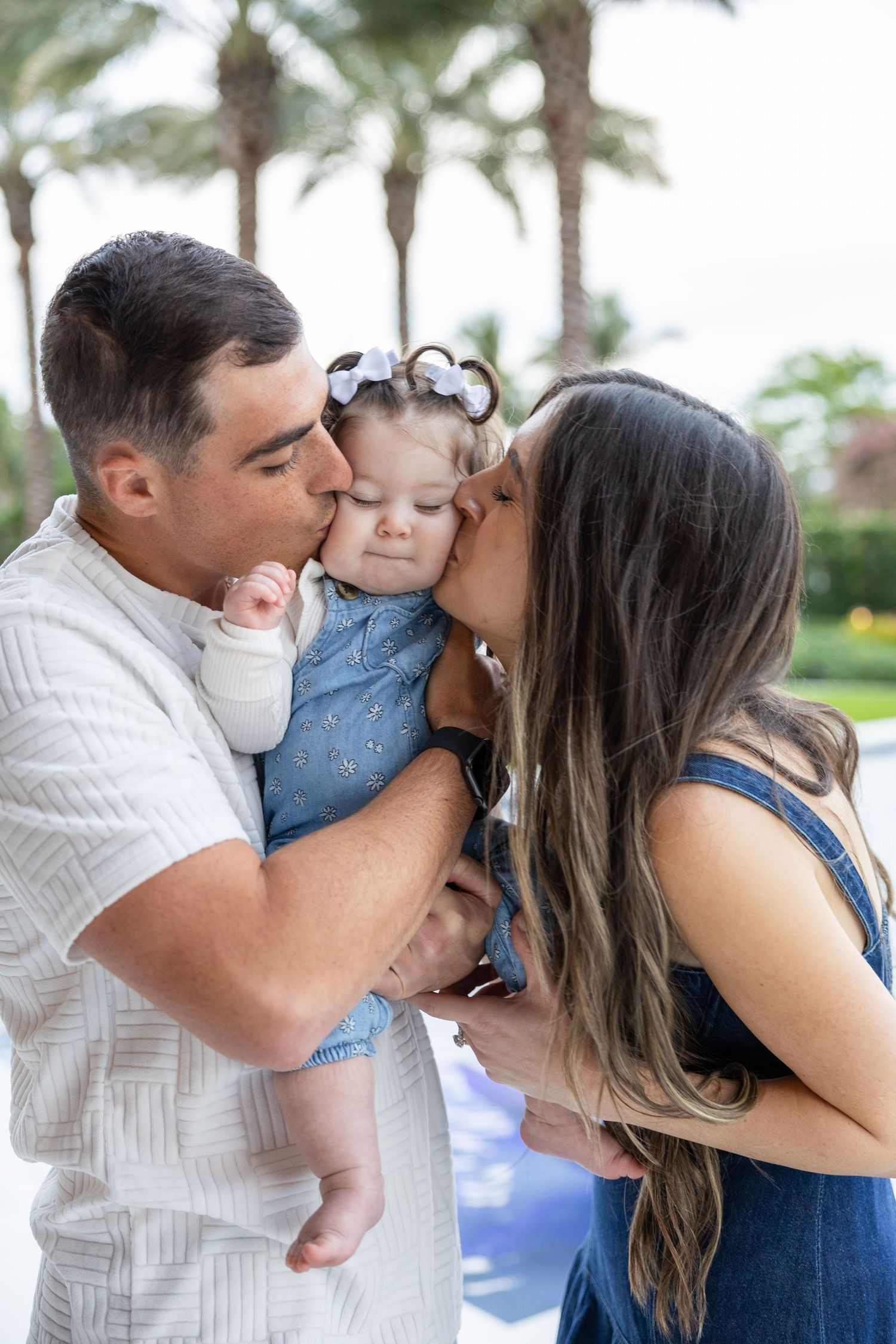 Parents kissing baby girl on cheeks outdoors with palm trees in background.
