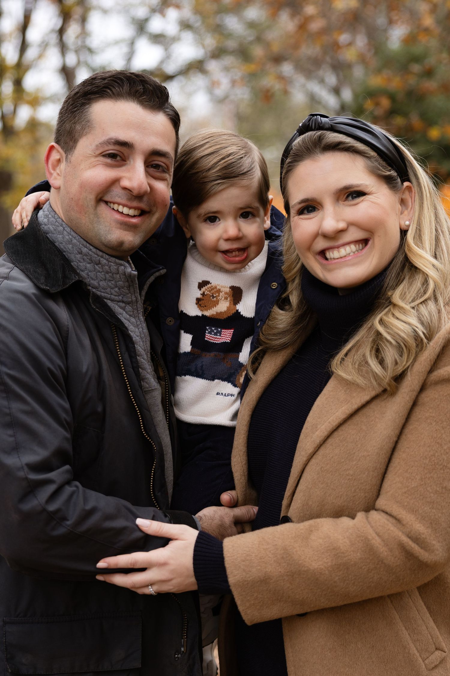 Happy family of three posing outdoors in fall, parents holding toddler boy in Ralph Lauren sweater.