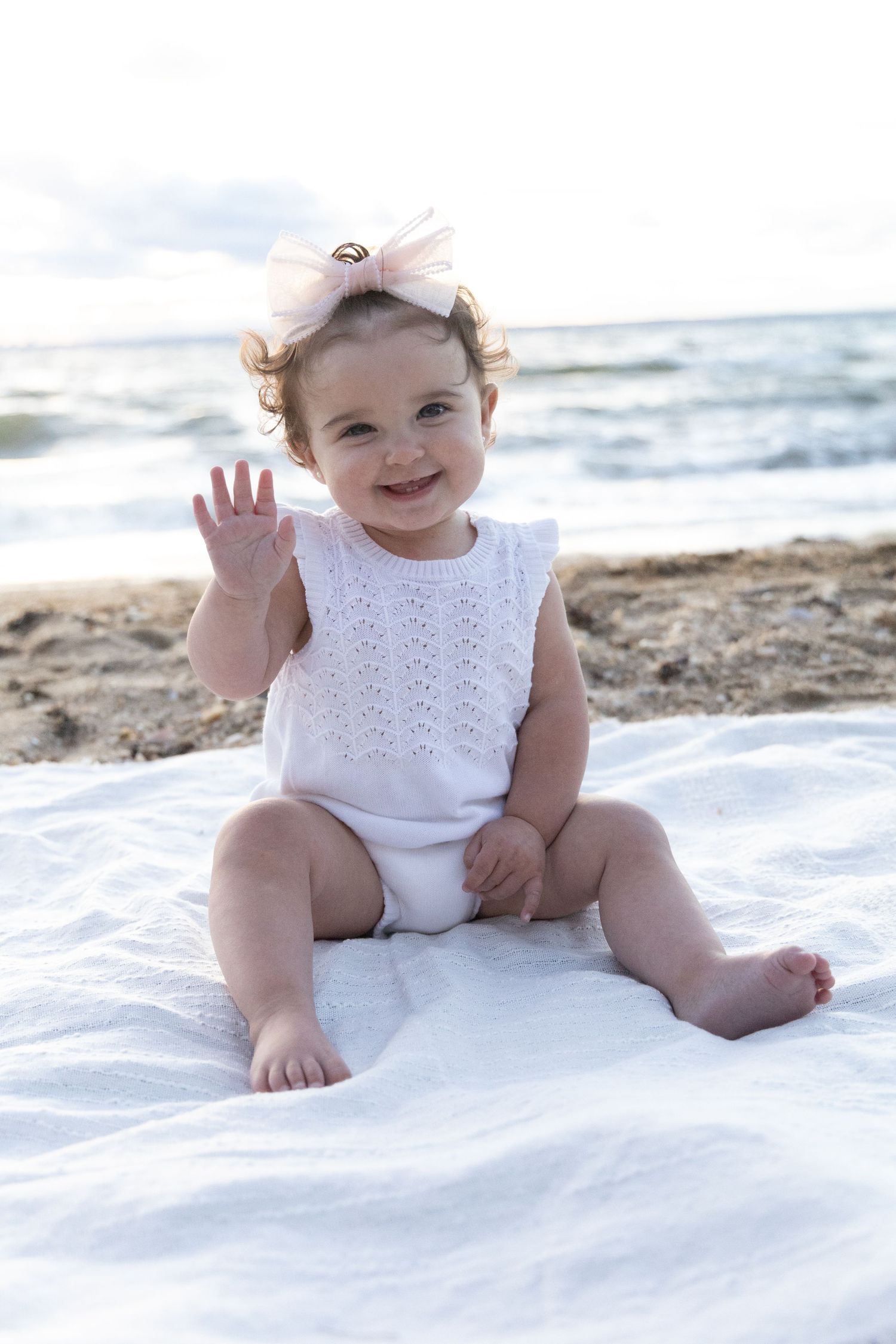 Smiling baby girl in white romper and pink bow headband waving on a white blanket at the beach.