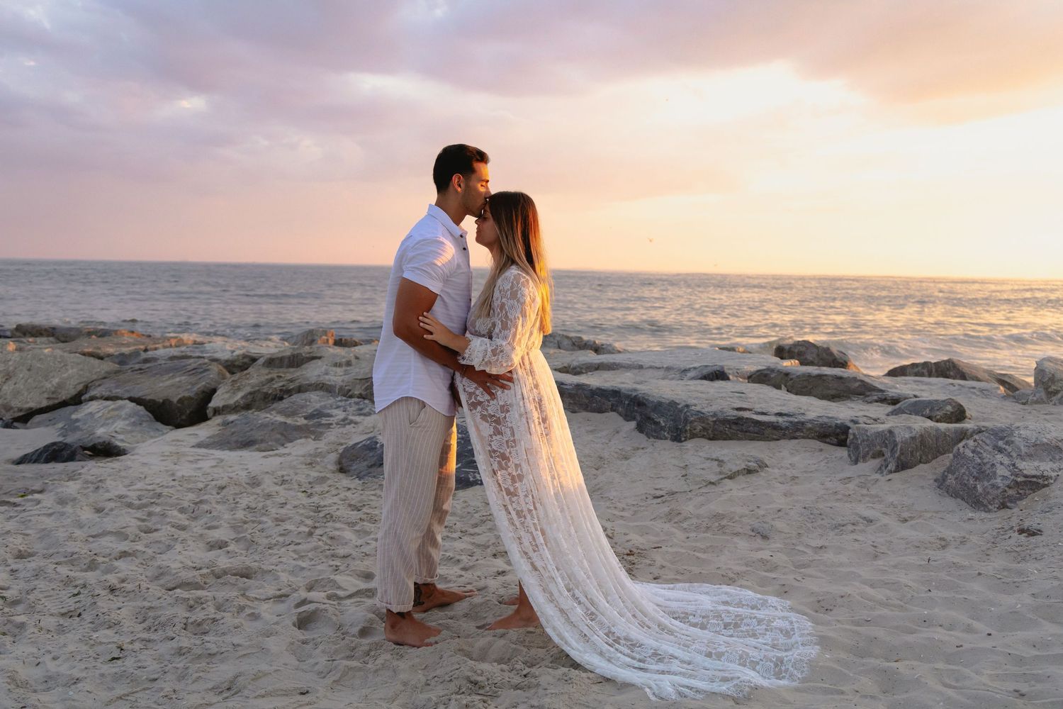 Couple sharing a romantic kiss at sunset on a rocky beach, woman in white lace gown.