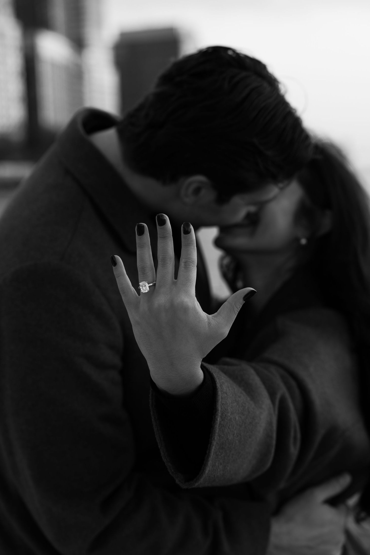 Black and white engagement photo of couple kissing with bride showing large diamond ring to camera.