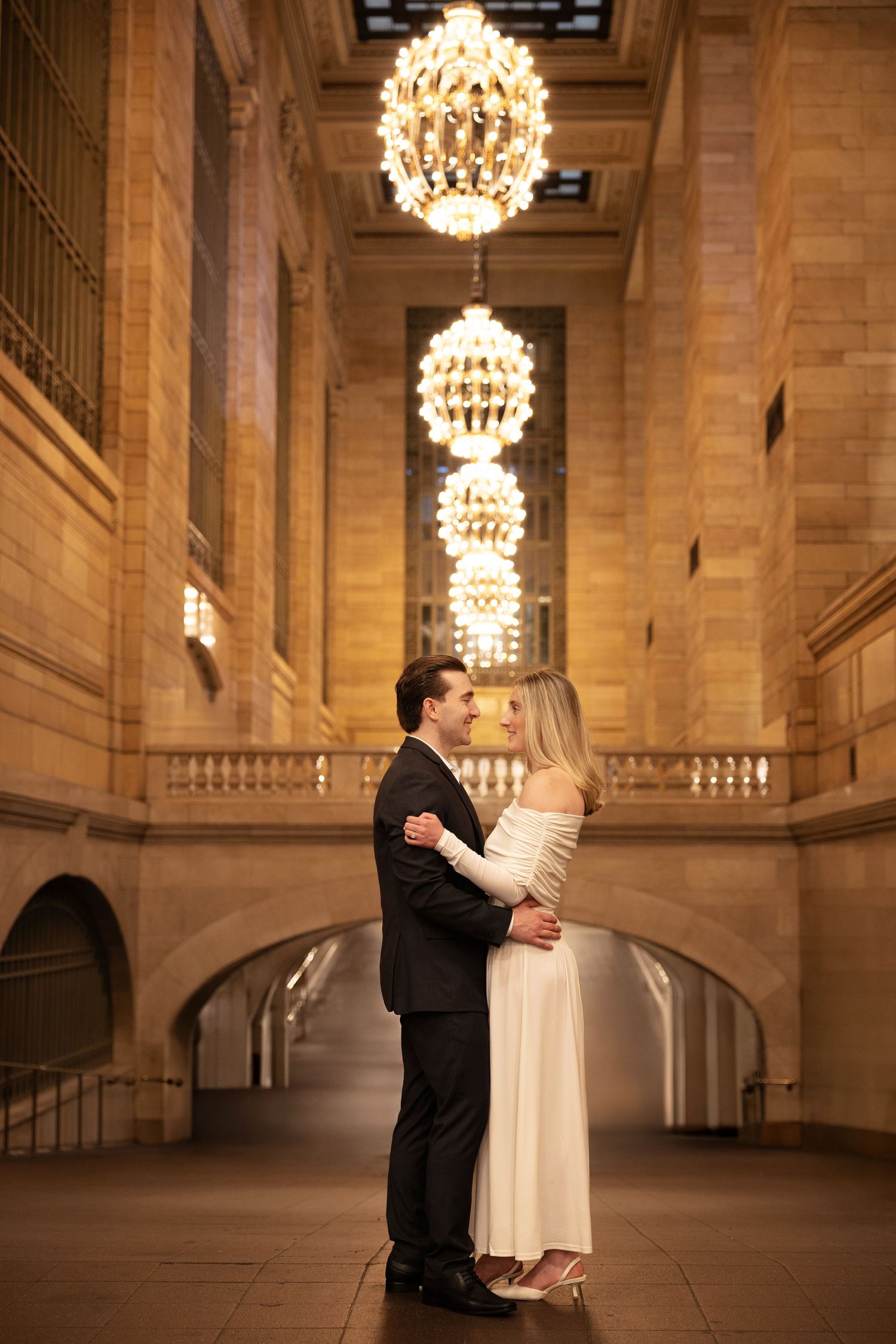 Romantic couple in formal attire embracing beneath glowing chandeliers in Grand Central Terminal NYC.
