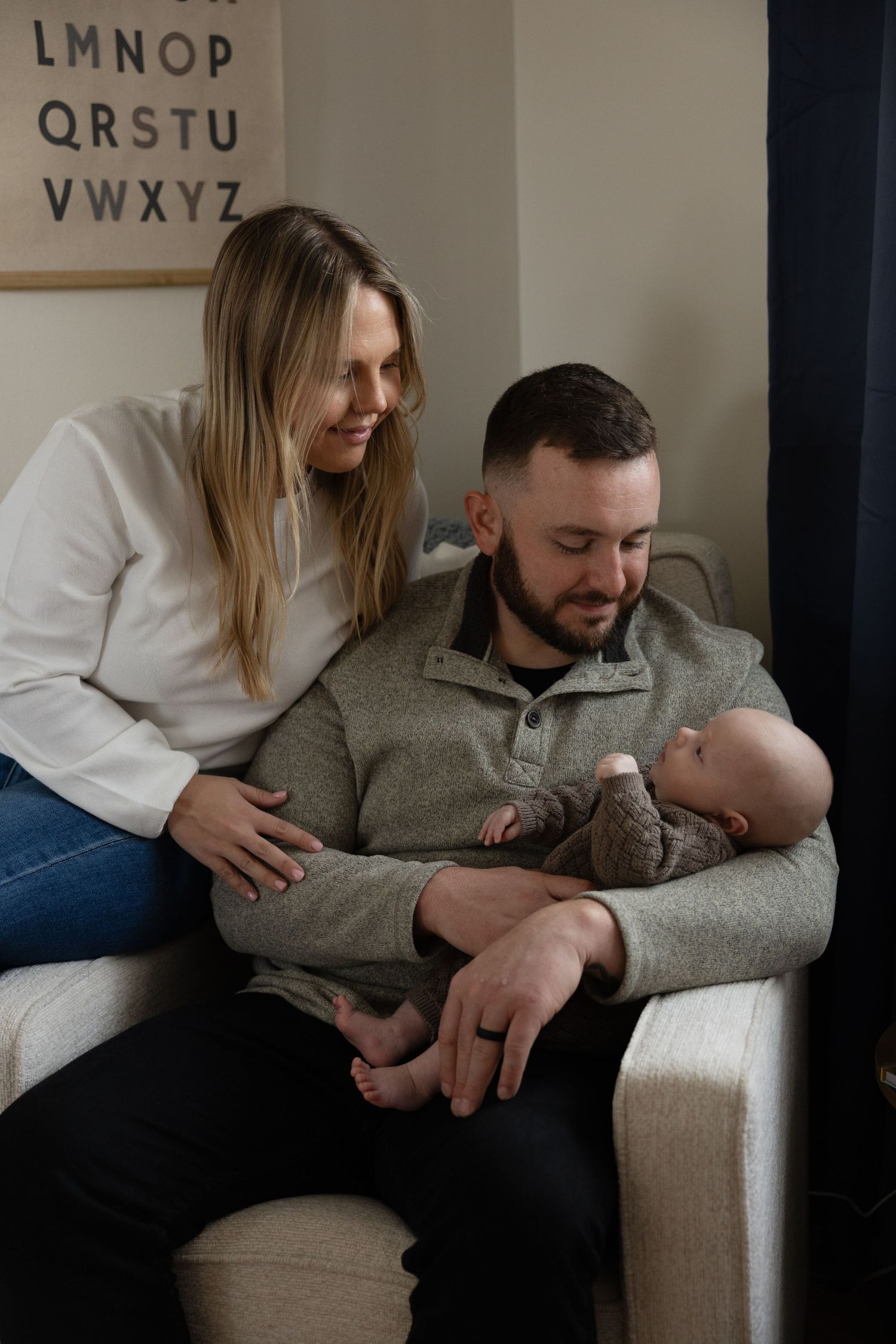 A couple smiles at their newborn baby, with the father cradling the infant in a cozy nursery chair.