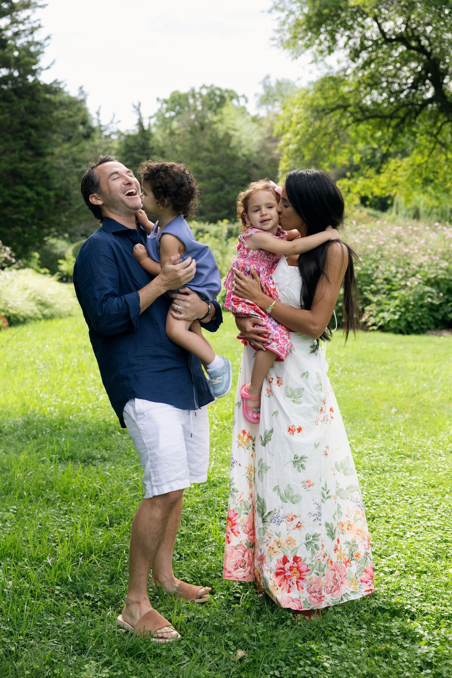 Happy family of four enjoying a sunny day in a lush green park, parents kissing their young children.