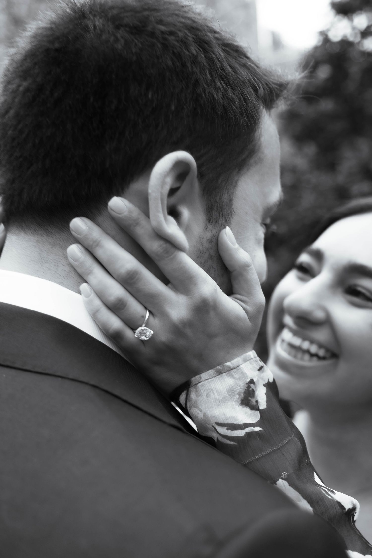 Black and white close-up of smiling bride showing diamond engagement ring while embracing groom.