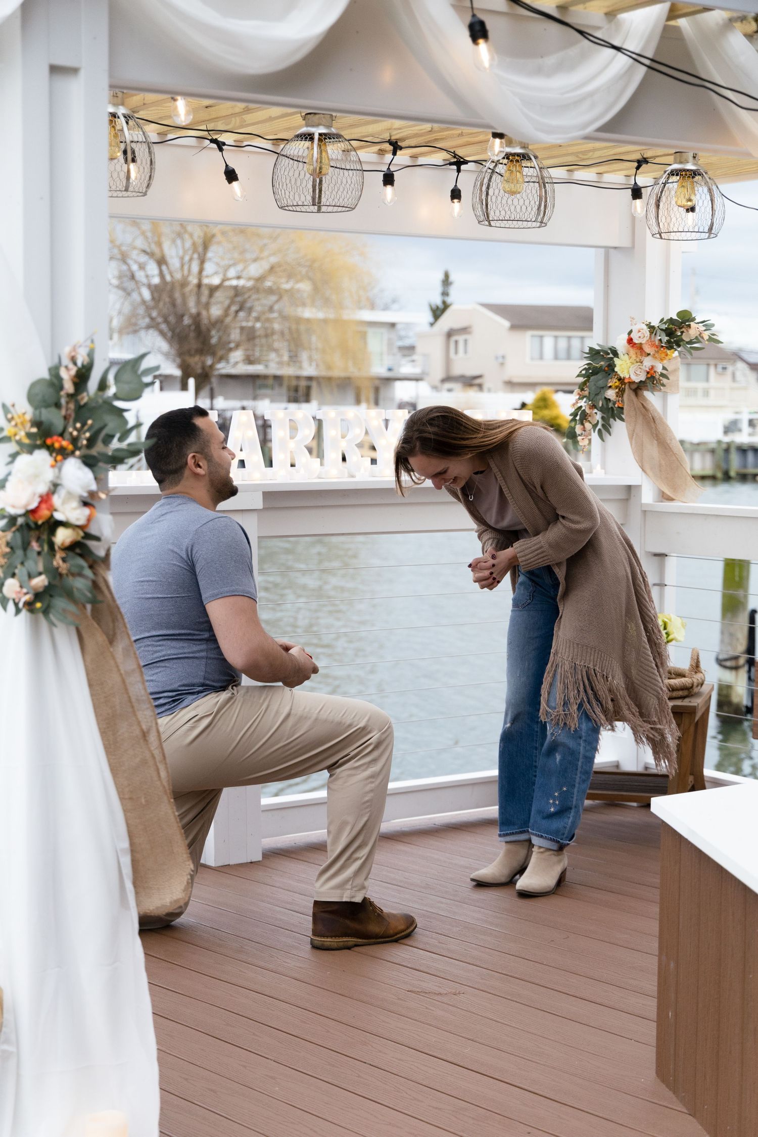 Man proposing on one knee to woman on decorated waterfront deck with floral arrangements and string lights.