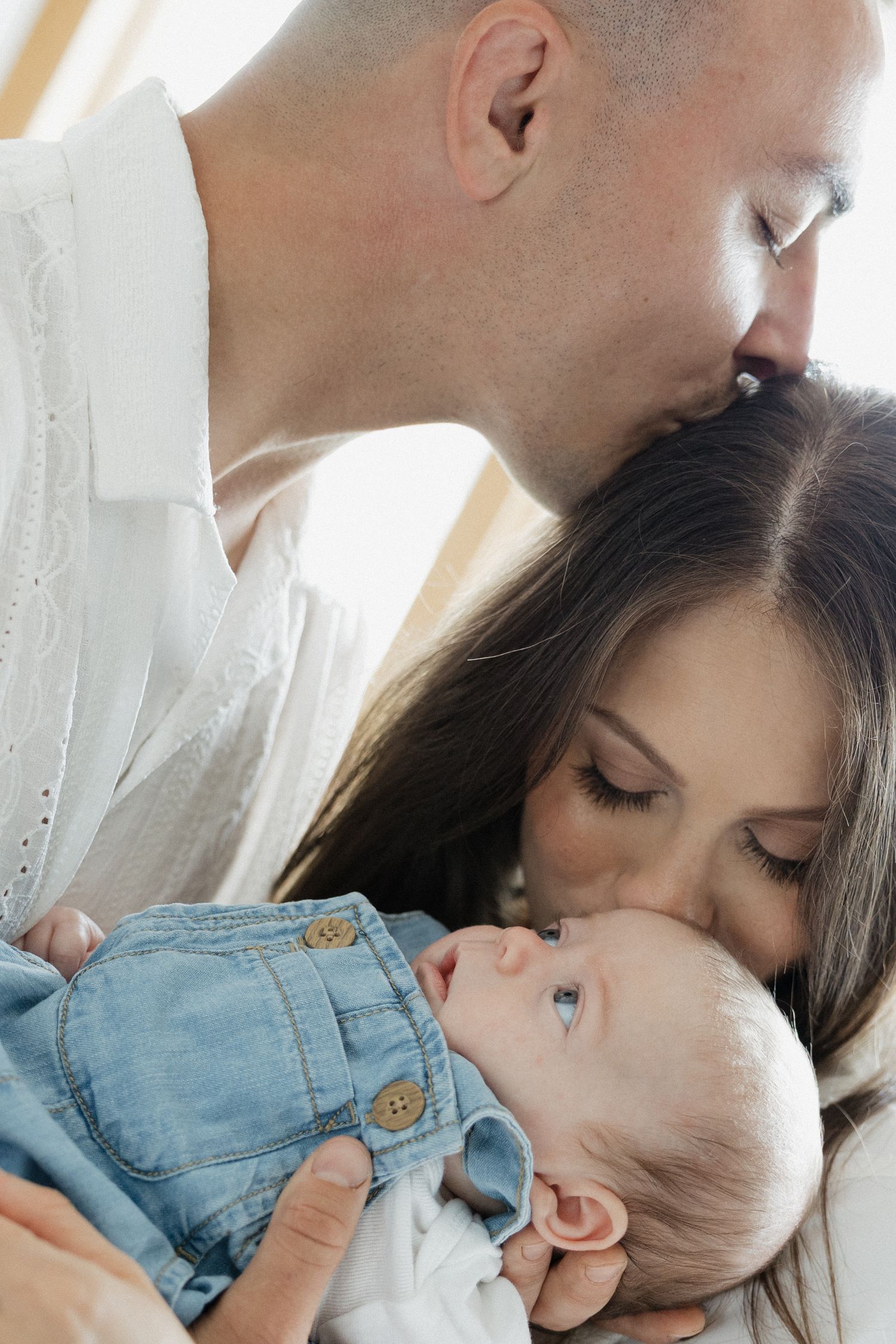 Tender moment of parents kissing while lovingly holding their wide-eyed newborn baby in white setting.