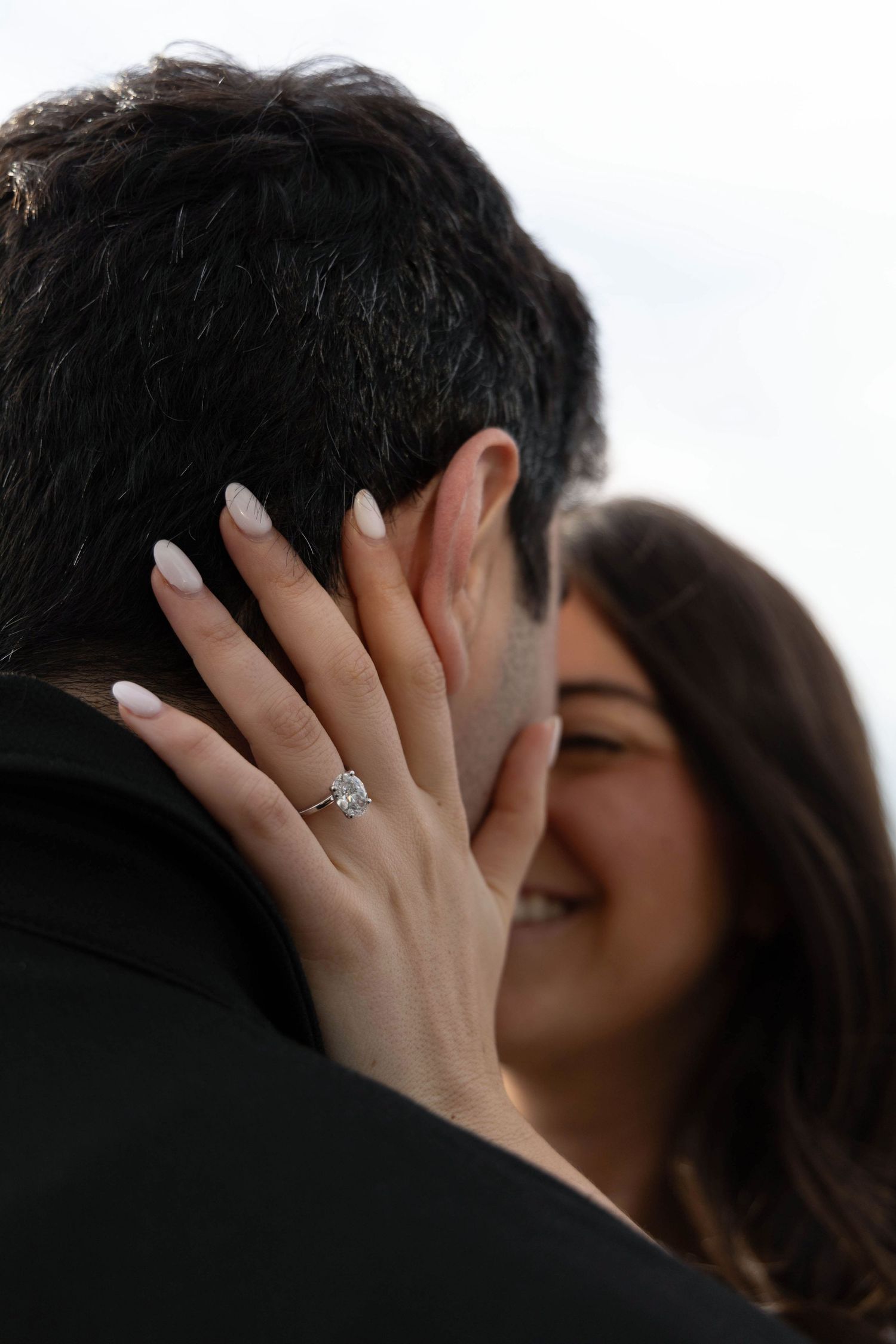 Close-up of womans hand with diamond engagement ring resting on mans face during romantic moment.