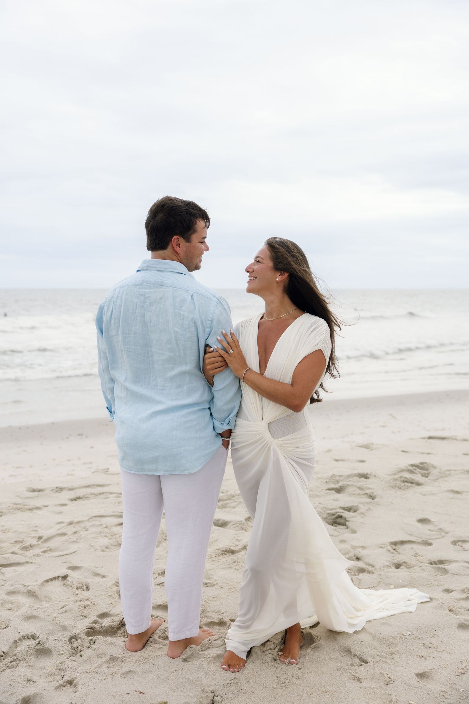Happy couple in white and blue outfits embracing on a sandy beach with ocean waves in the background.