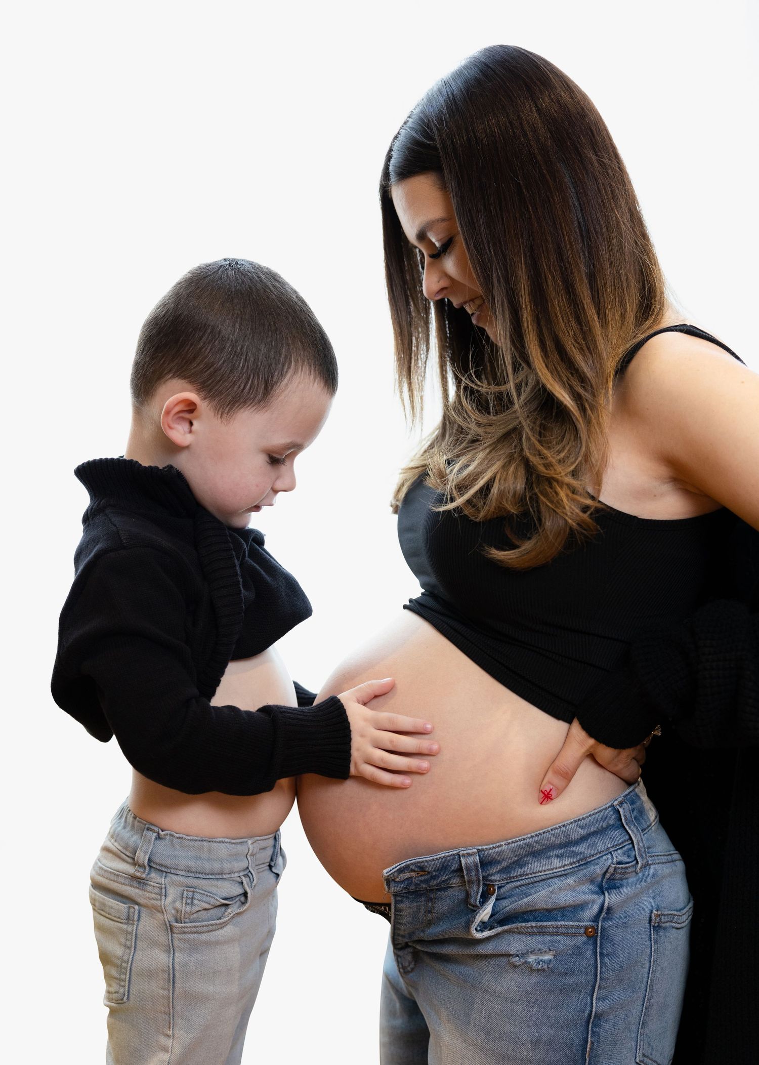 Young boy touching pregnant mothers bare belly during maternity photo session on white background.