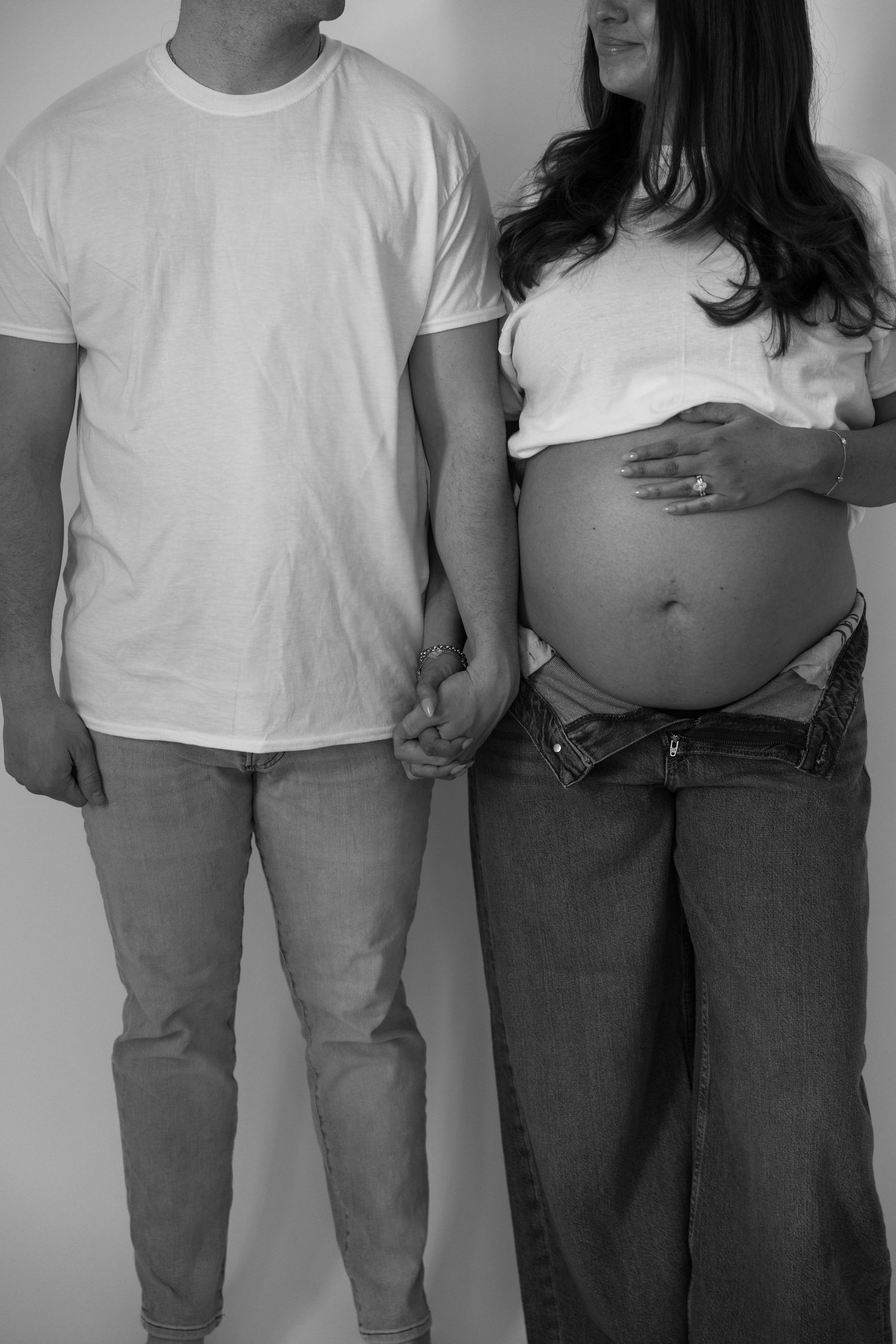Black and white photo of pregnant couple holding hands, woman cradling baby bump in white top and jeans.