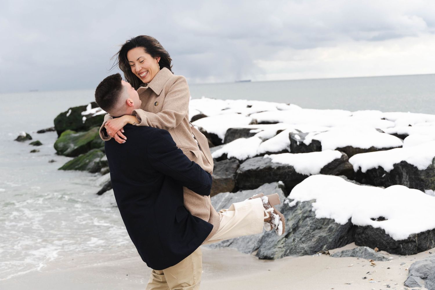 Man lifting woman on a snowy beach with snow-covered rocks and ocean waves in the background.