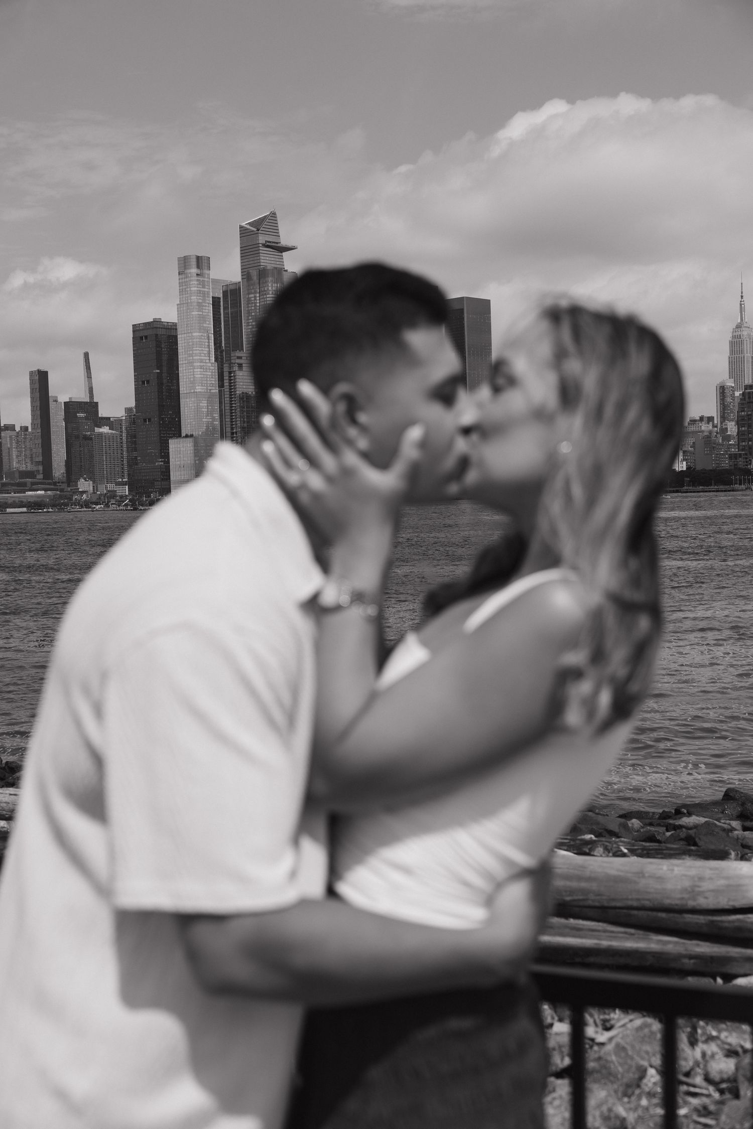 Black and white photo of couple kissing with New York City skyline and Hudson River in background.