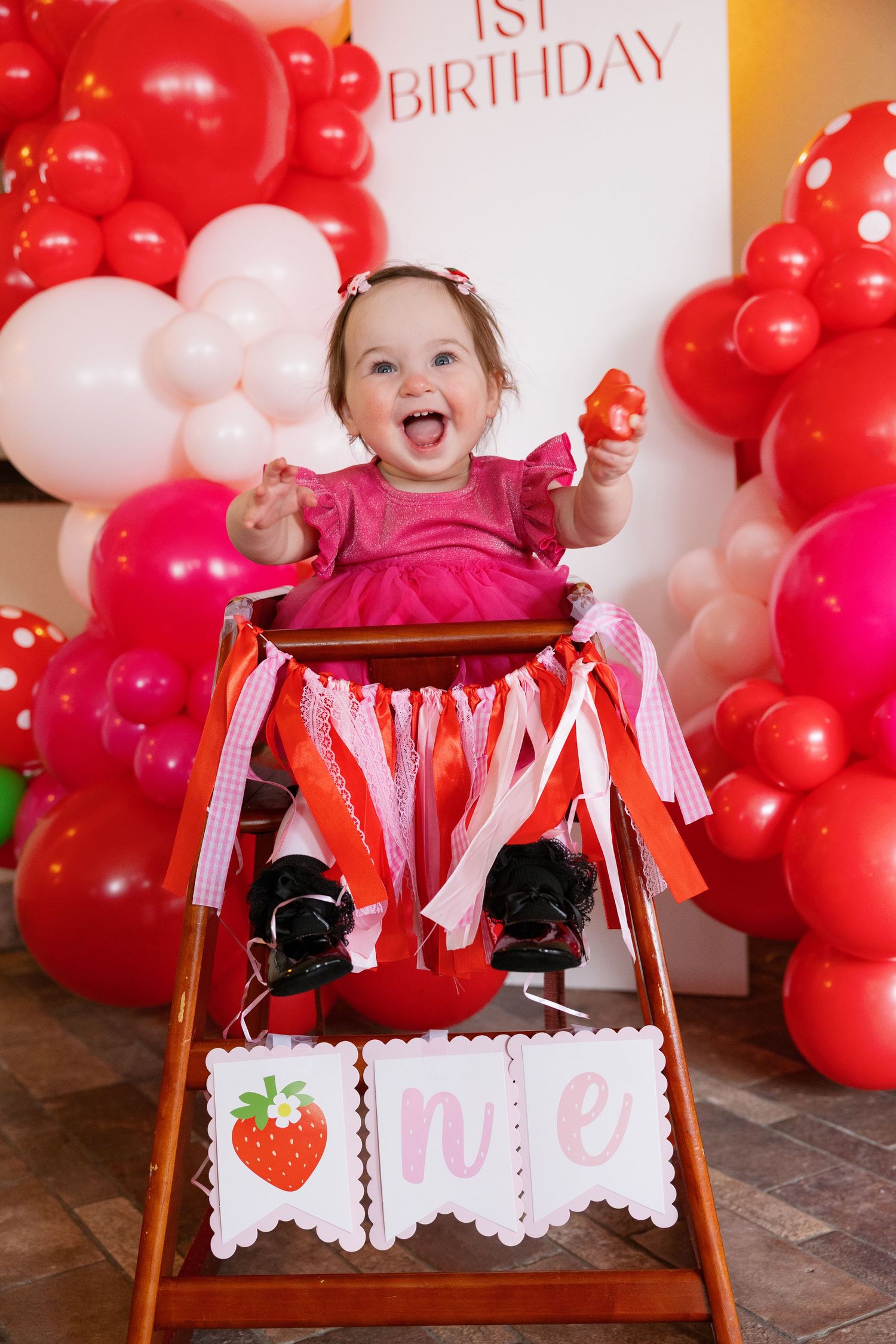 Happy baby girl in pink dress celebrating her first strawberry-themed birthday in a high chair.