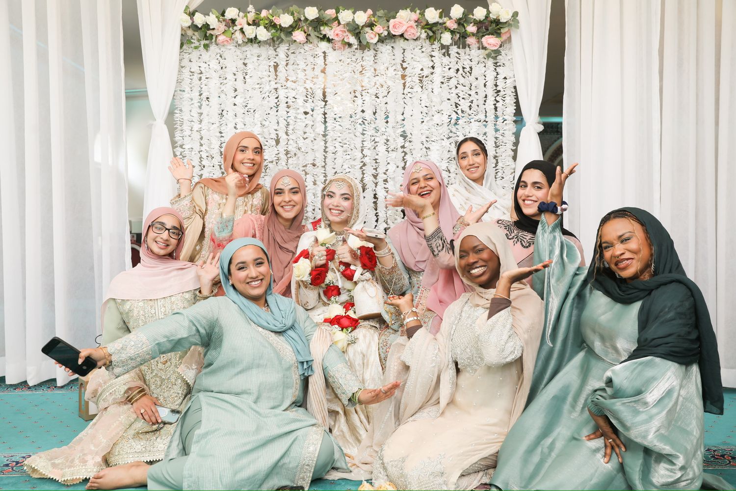 A group in pastel and mint colored traditional attire poses together in front of a floral backdrop with string lights.