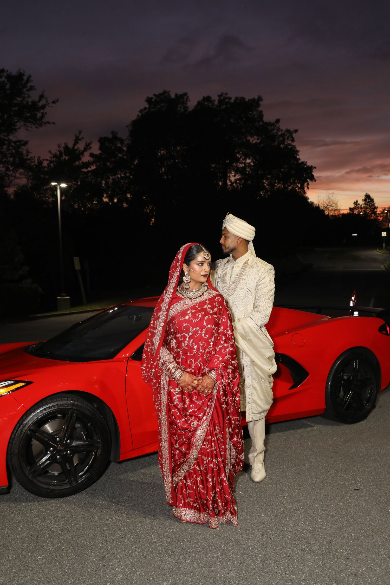 Traditional South Asian wedding couple posing with a bright red Ferrari sports car in a parking lot.