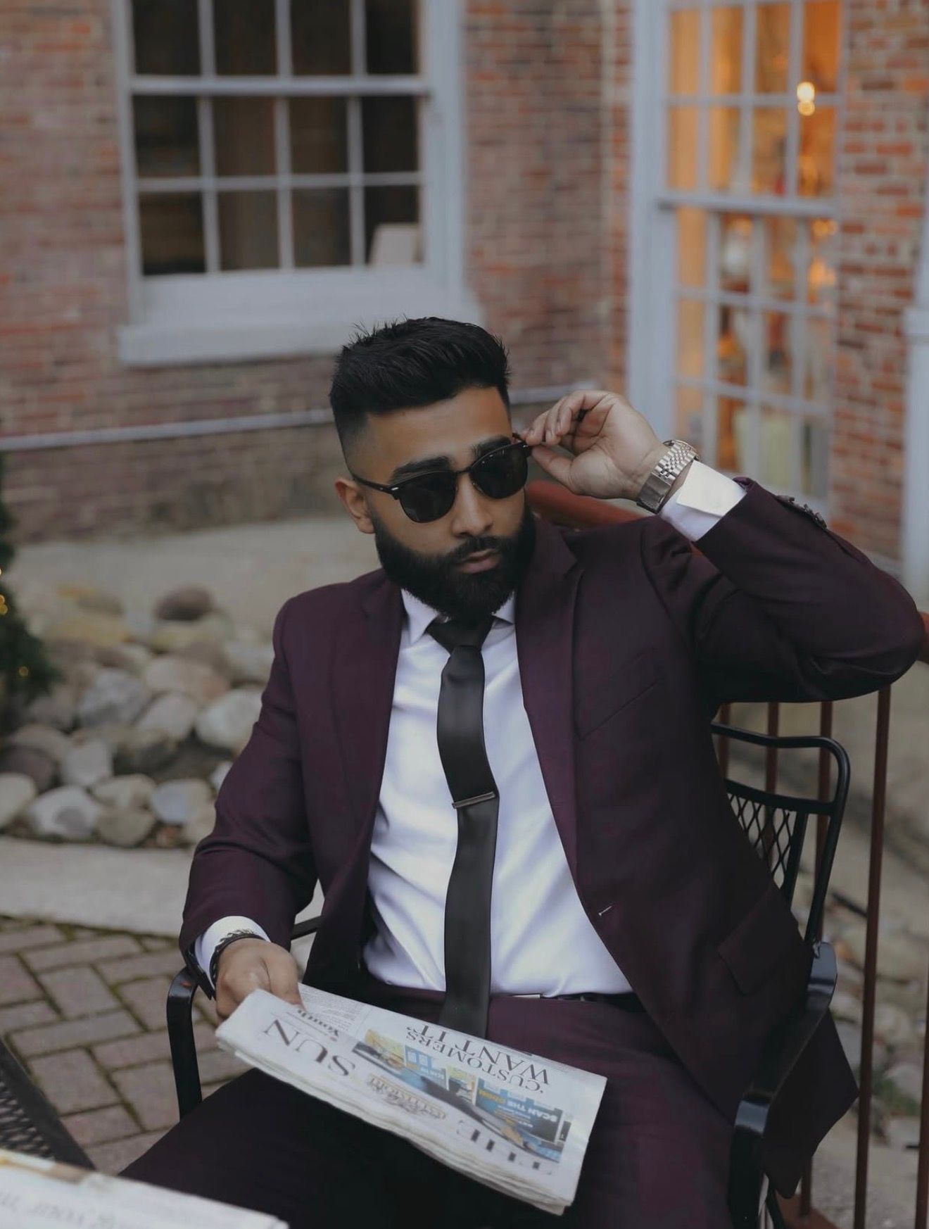 A person in a burgundy suit and sunglasses sits in a chair reading a newspaper in an outdoor brick setting.