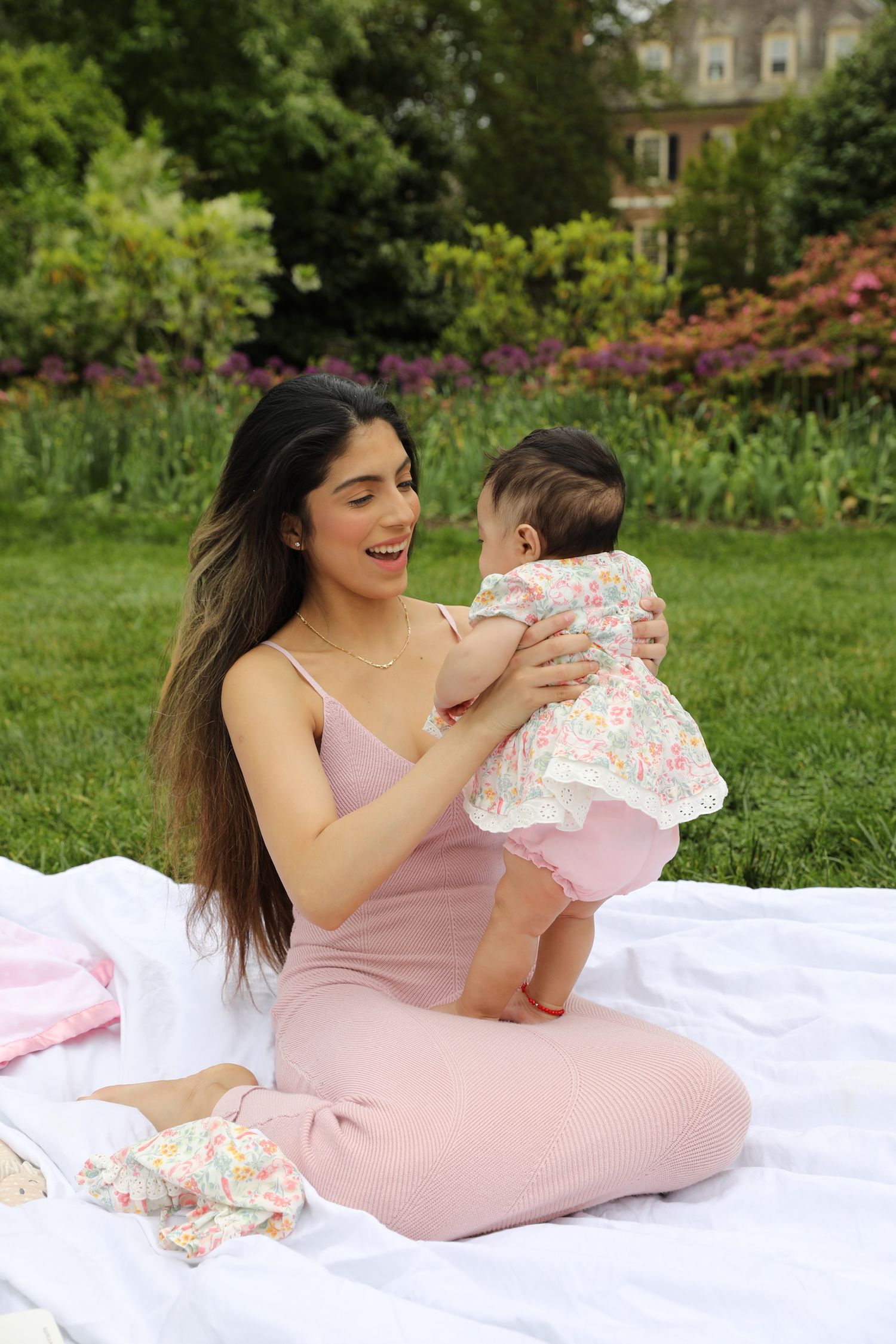 A parent and baby share a tender moment on a pink blanket in a lush garden setting with blooming flowers.