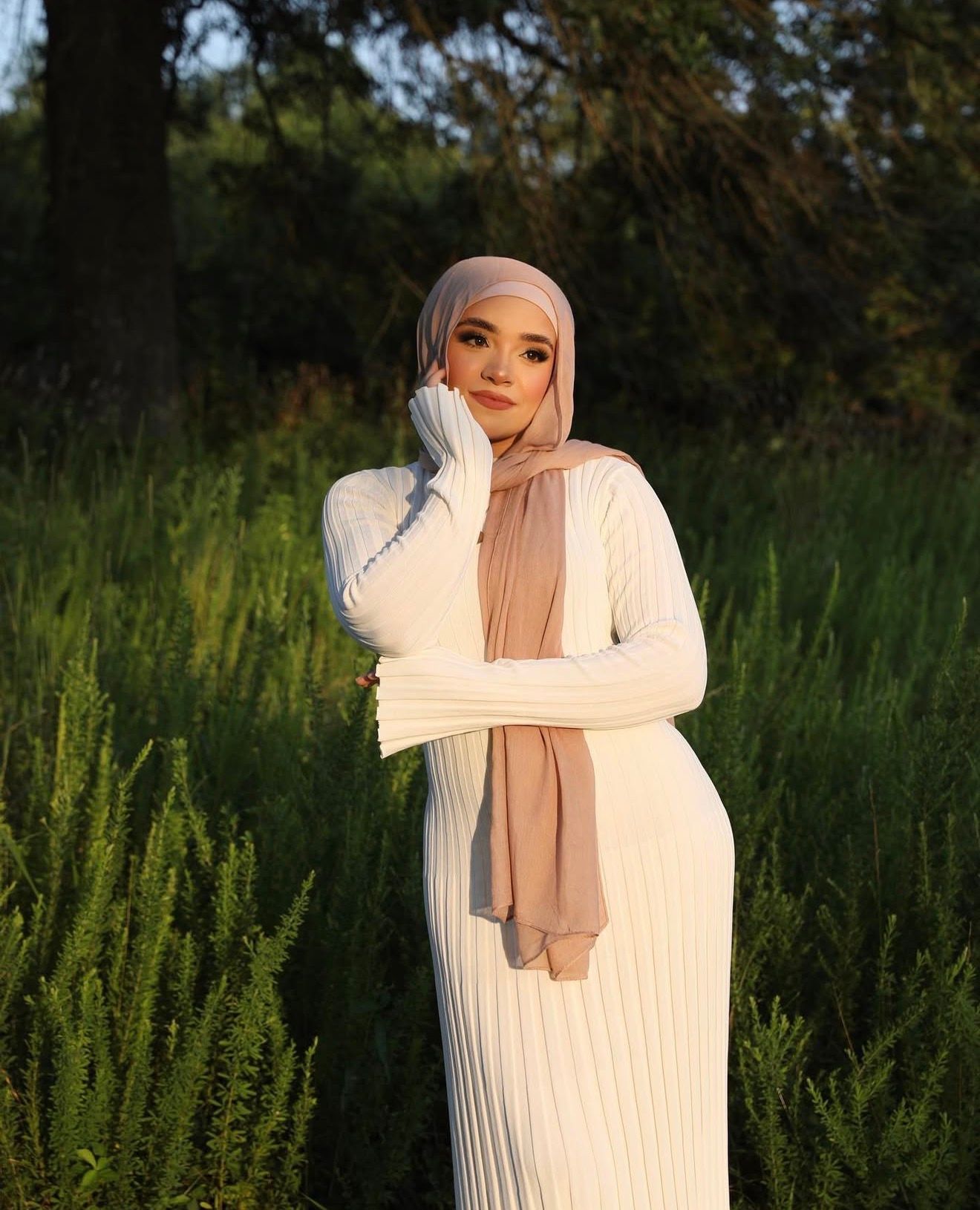 Person in white sweater and light headscarf poses outdoors in golden hour lighting.