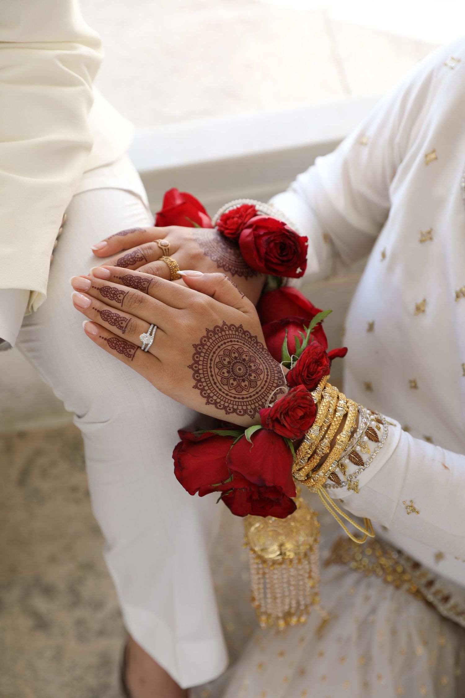 Close-up of adorned hands with henna design, red roses, and gold bangles against white traditional attire.