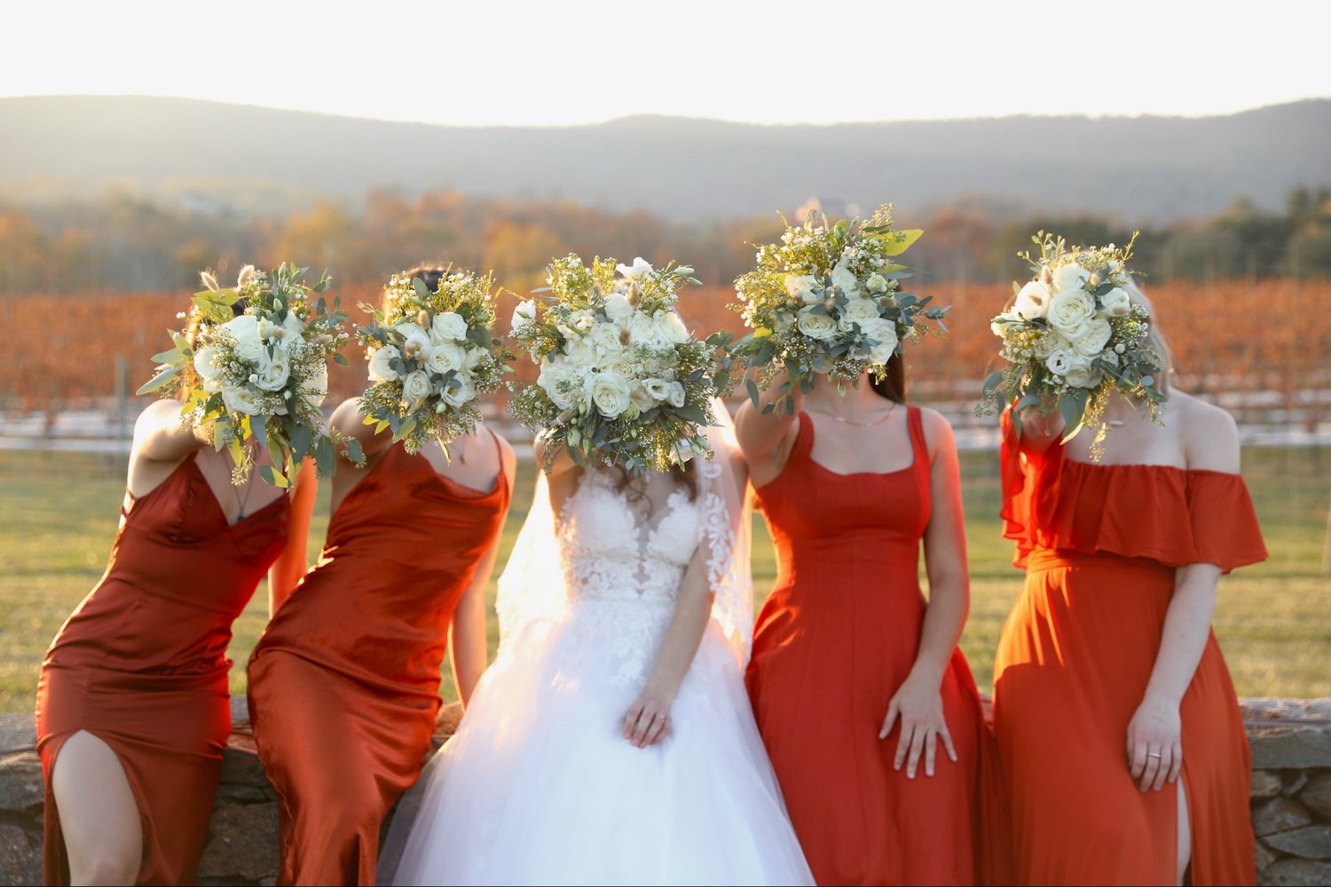 Wedding party in matching burnt orange dresses poses together outdoors with white floral bouquets.