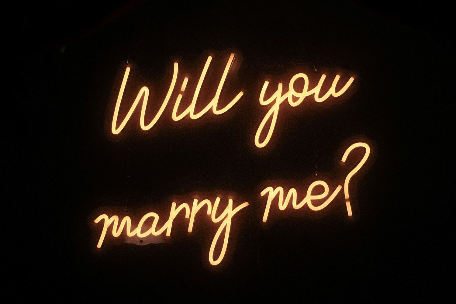 Glowing neon sign displaying 'Will you marry me?' in cursive yellow lettering against a dark background.