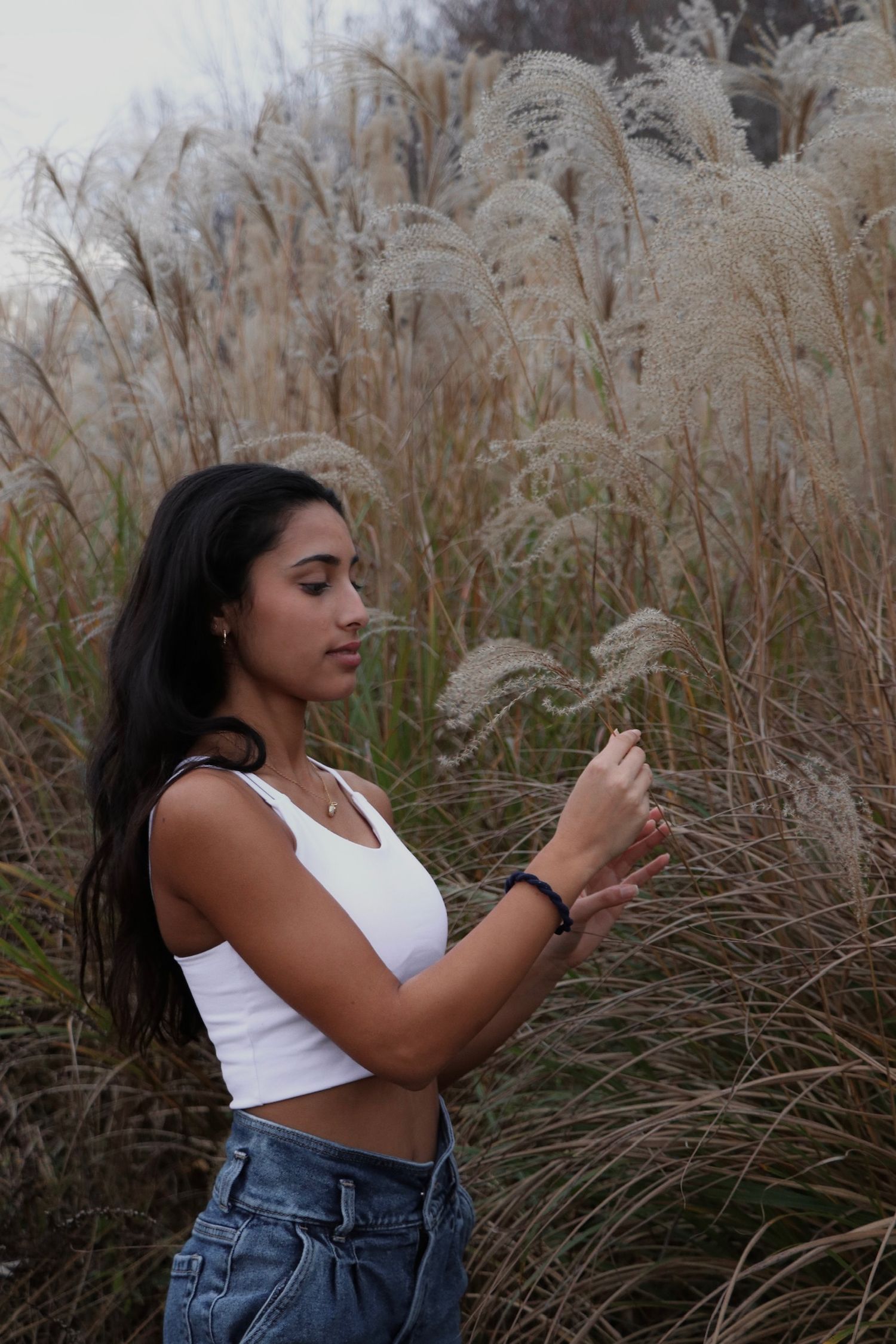 A person in a white crop top and jeans poses against tall pampas grass in a moody outdoor setting.