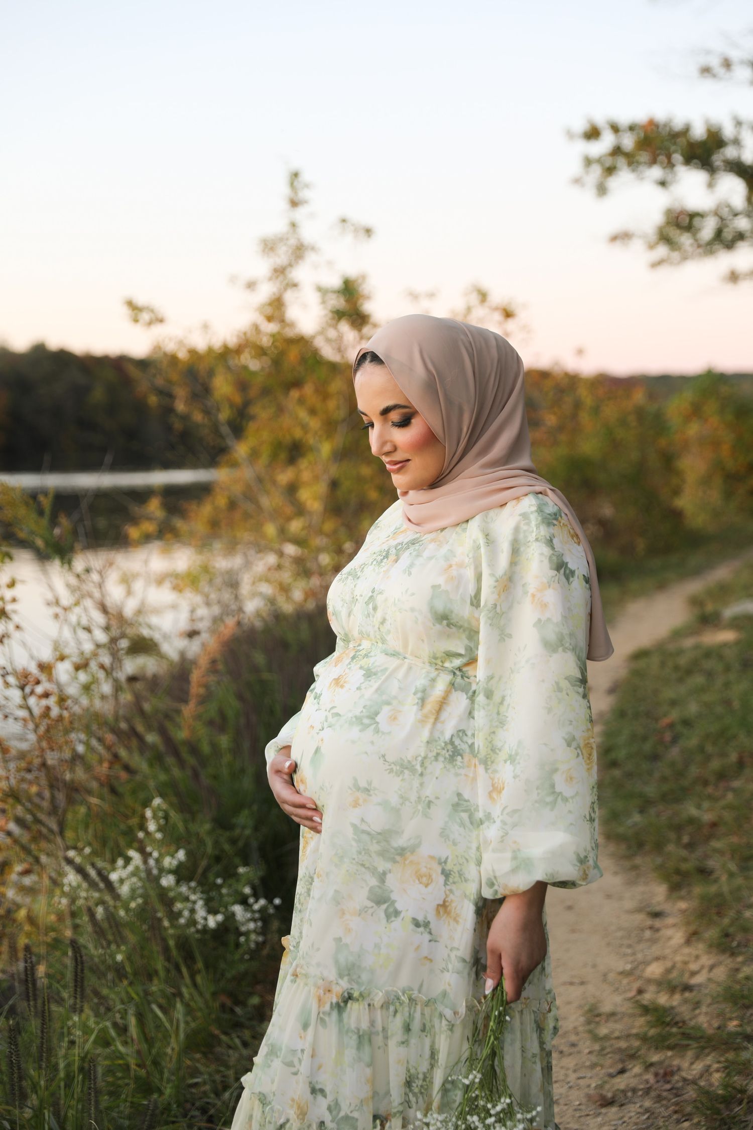 Woman in floral dress and hijab poses along a scenic dirt path during golden hour sunset.