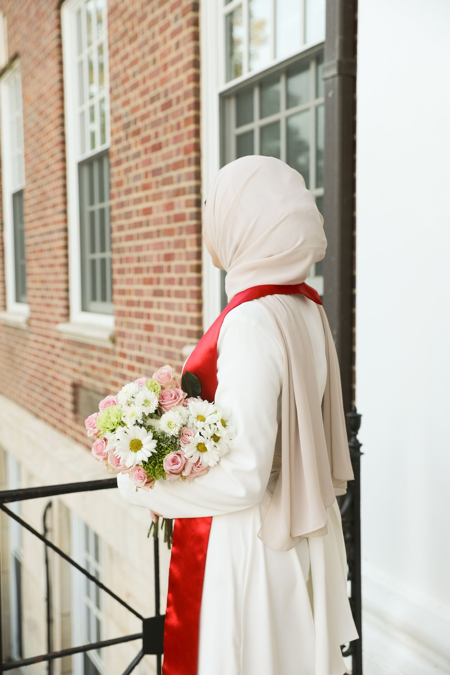 Person in white hijab and red accent dress holds a beautiful bouquet of pink and white flowers on brick building steps.