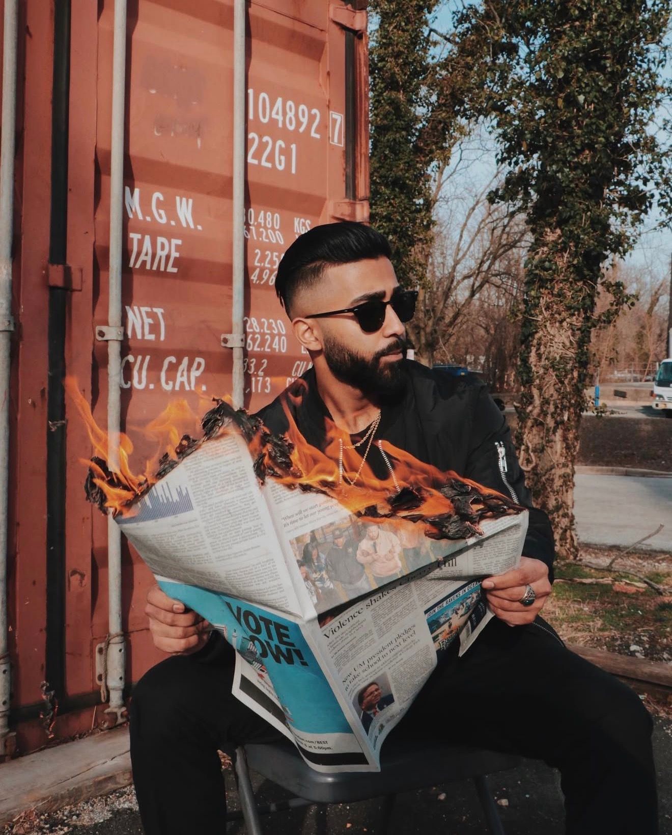 A person in sunglasses and black jacket reads a newspaper while sitting outdoors on a sunny day.