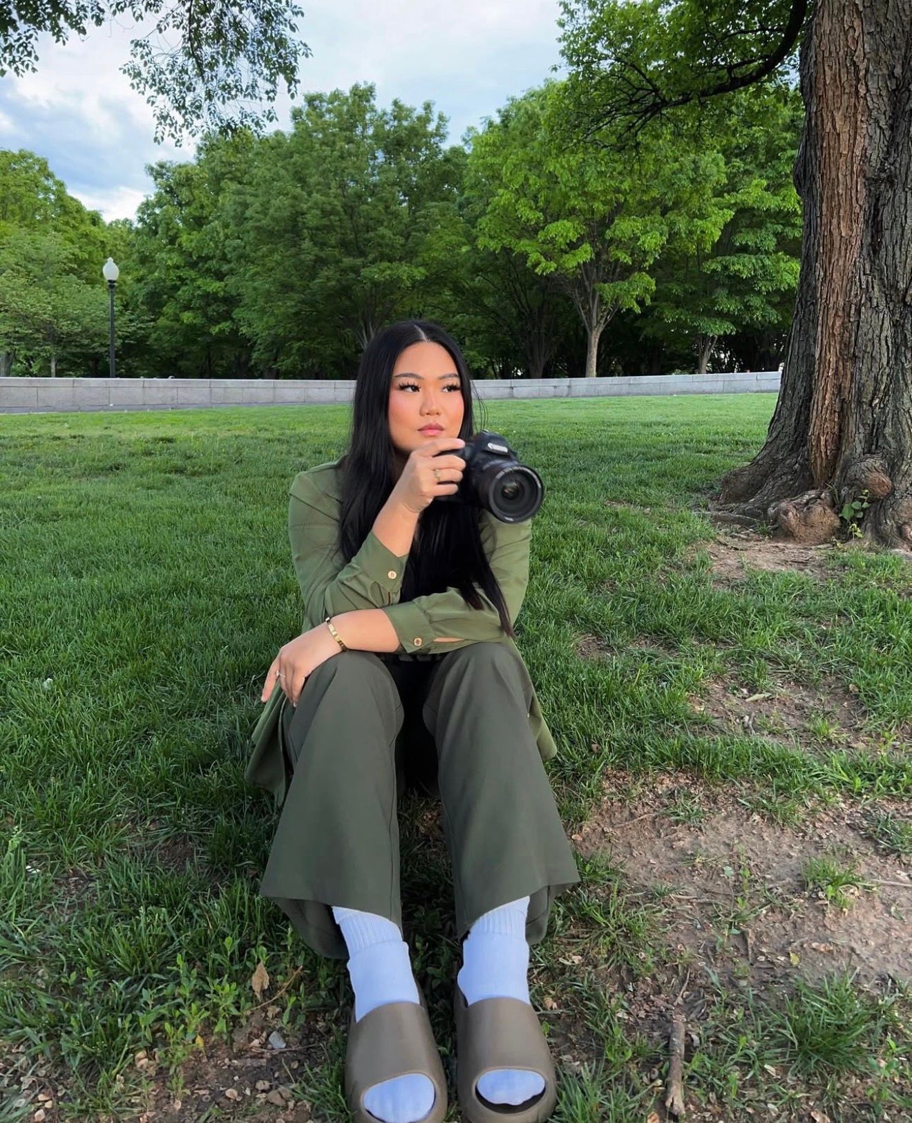 A photographer in olive green attire sits on grassy ground taking photos with a professional camera in a park setting.