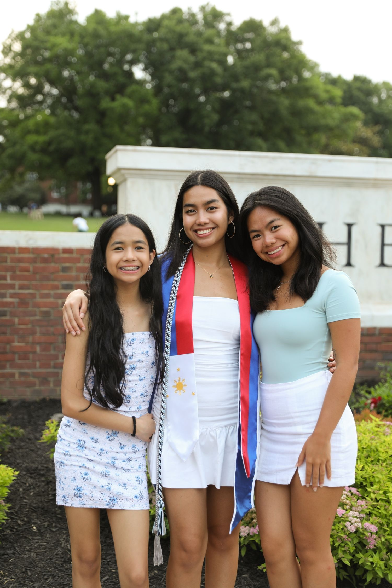 Three people in white summer outfits pose together outdoors near landscaped bushes and a brick wall.
