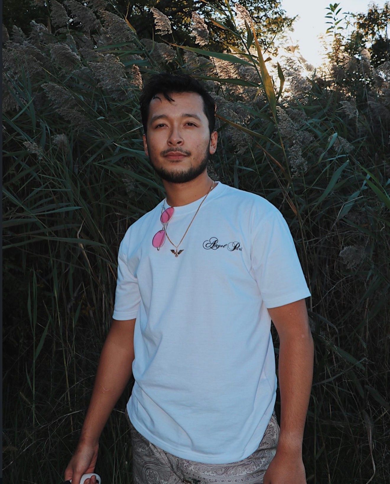 Person in white t-shirt standing outdoors against green foliage background at dusk.