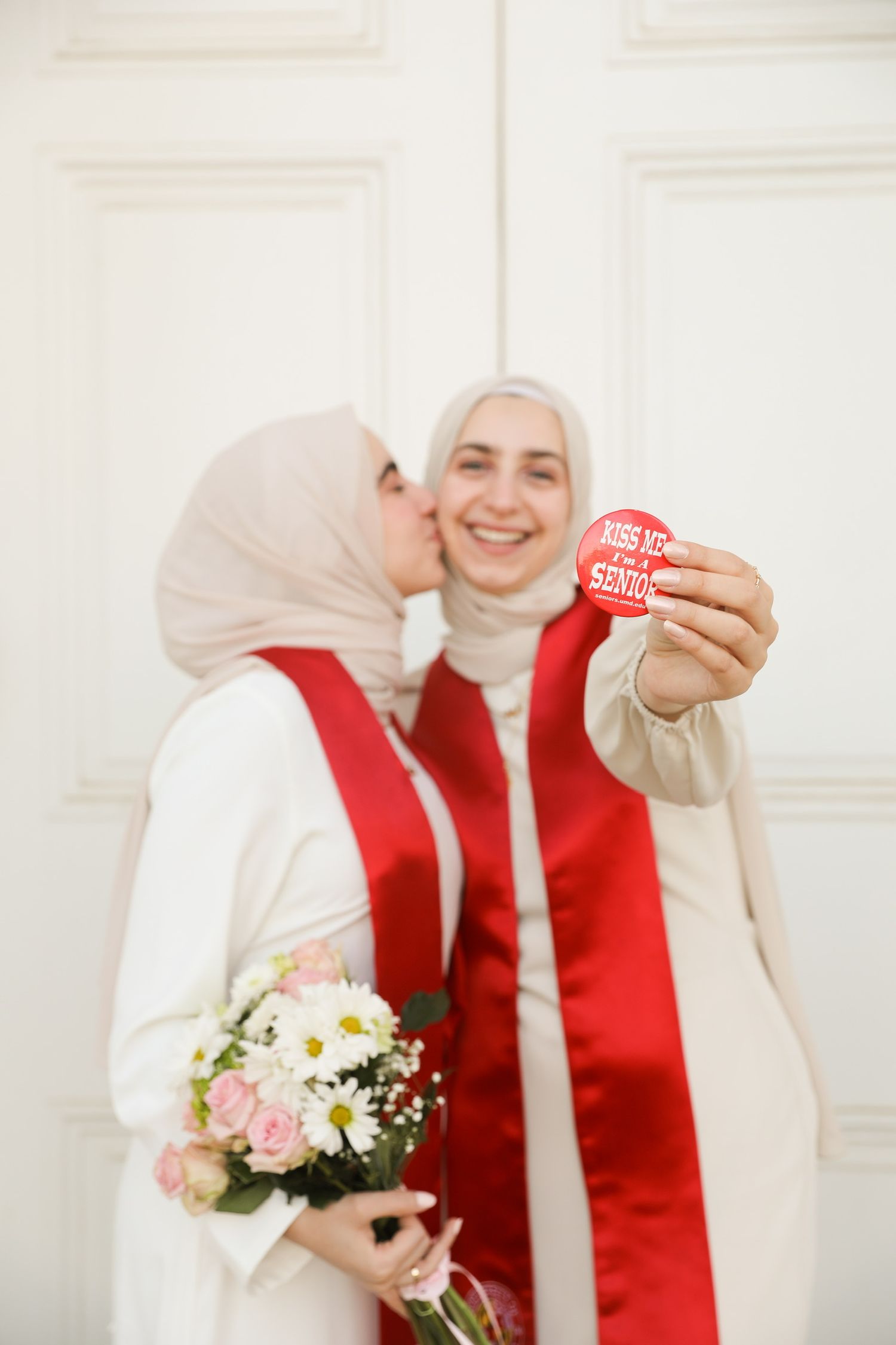 A person in a red vest and white headscarf holds a bouquet of pink and white flowers against a white background.
