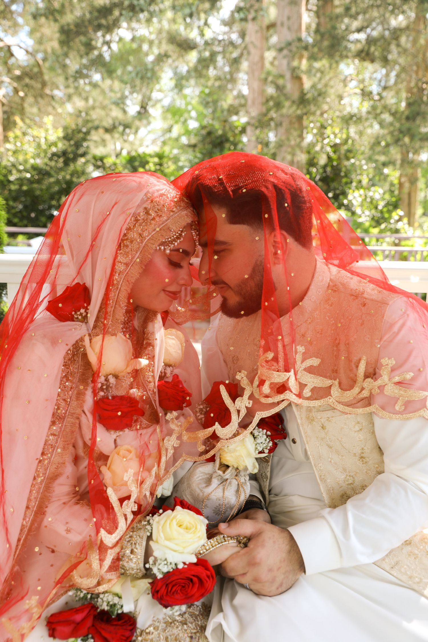 A romantic wedding couple shares tender moments under a flowing red veil in an outdoor garden setting.