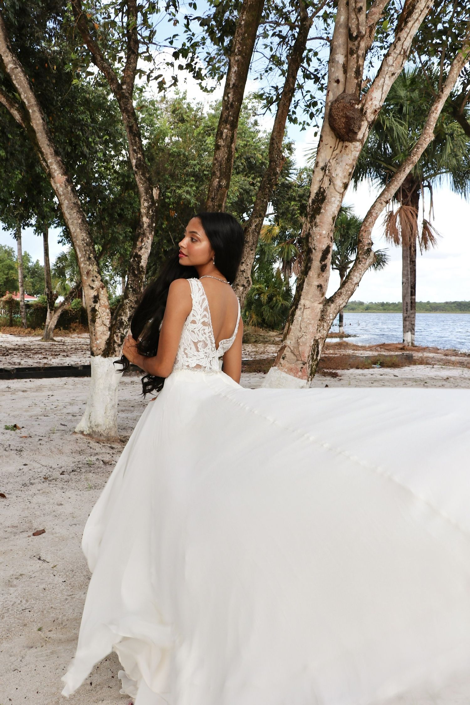A flowing white wedding dress billows in the breeze against a backdrop of palm trees and water at a beach location.