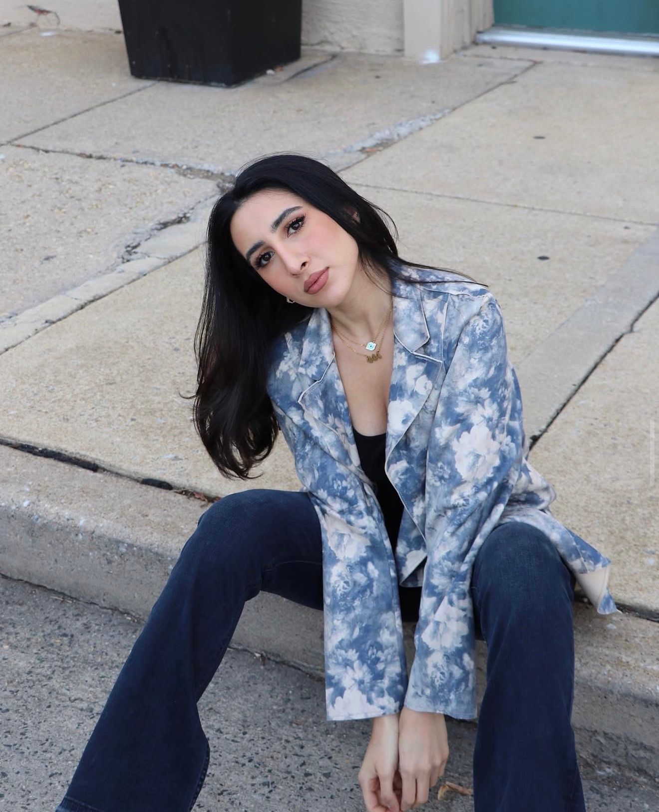 A person in a blue tie-dye blazer and dark pants poses on a concrete sidewalk in an urban setting.