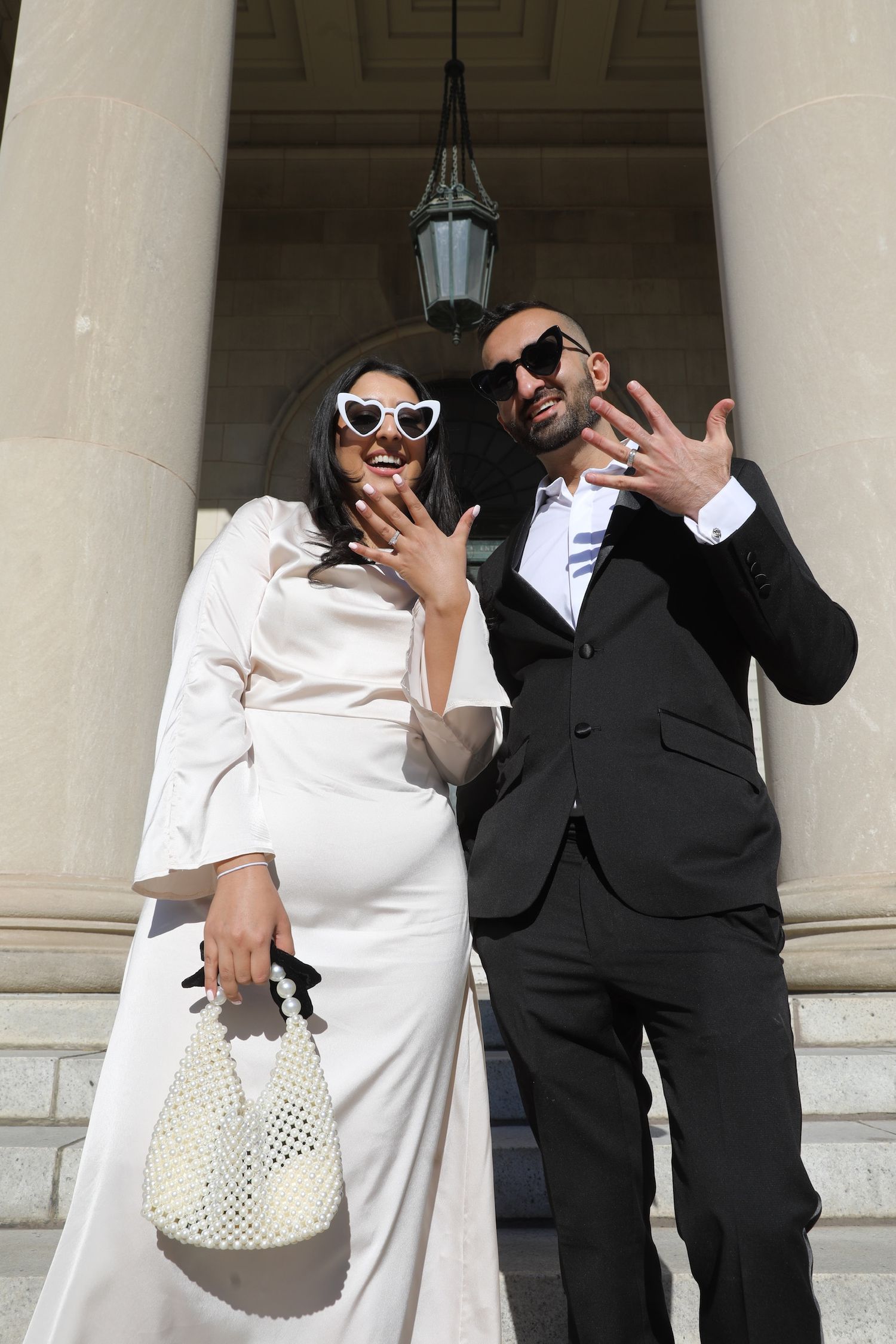 A couple in formal attire poses playfully in front of classical columns, with the person in white holding a woven bag.