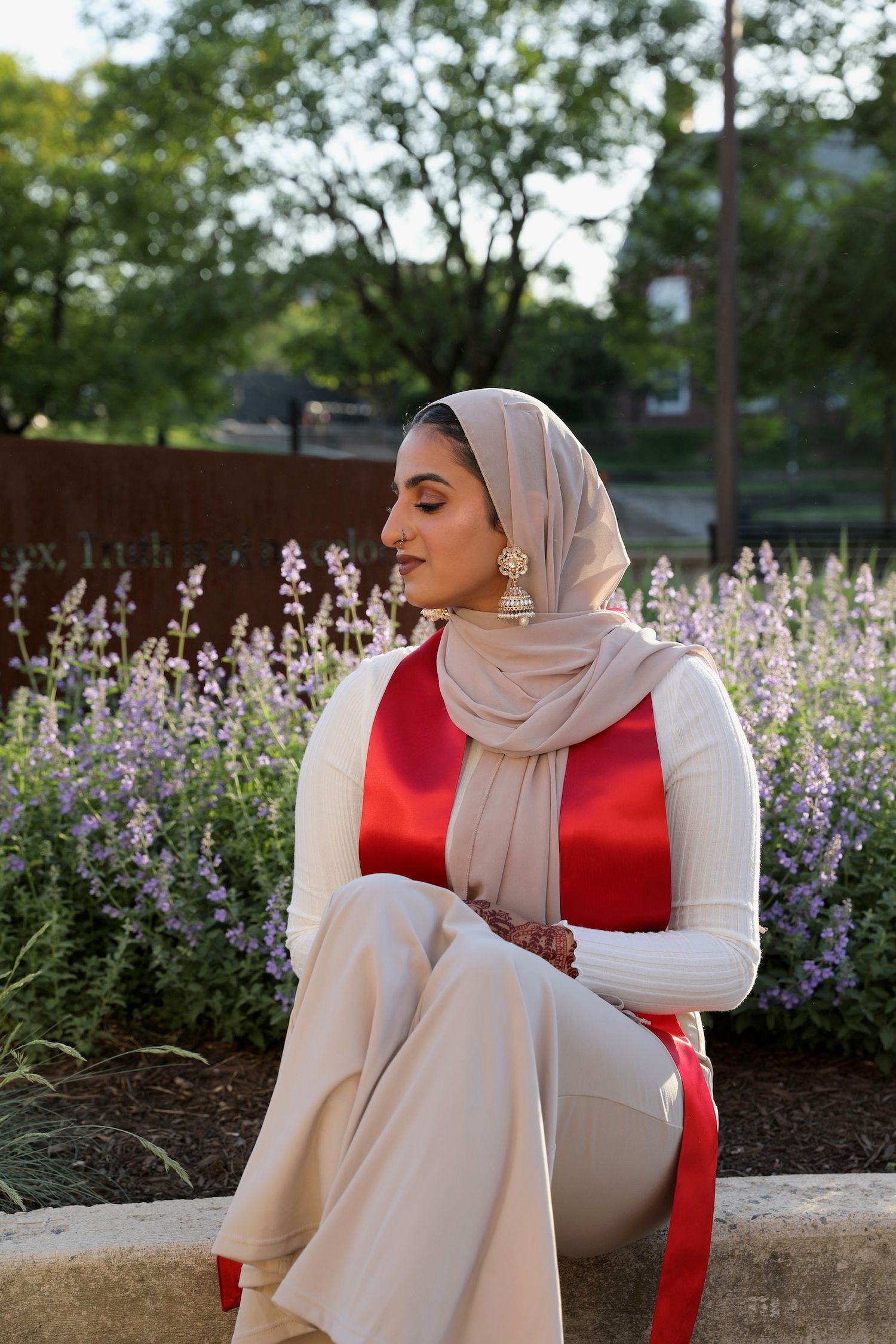 A person in a red top and beige hijab sits outdoors near blooming purple flowers in a garden setting.