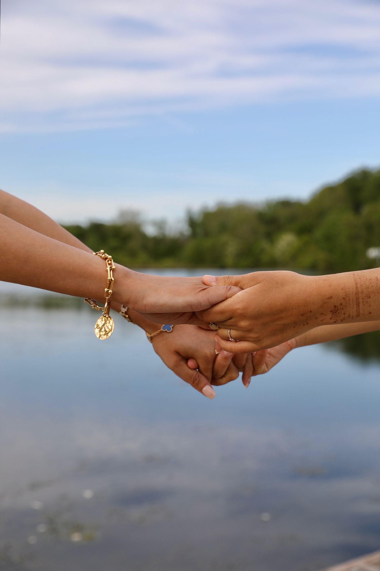 Two hands reach towards each other against a lake backdrop, one wearing a decorative gold ring.