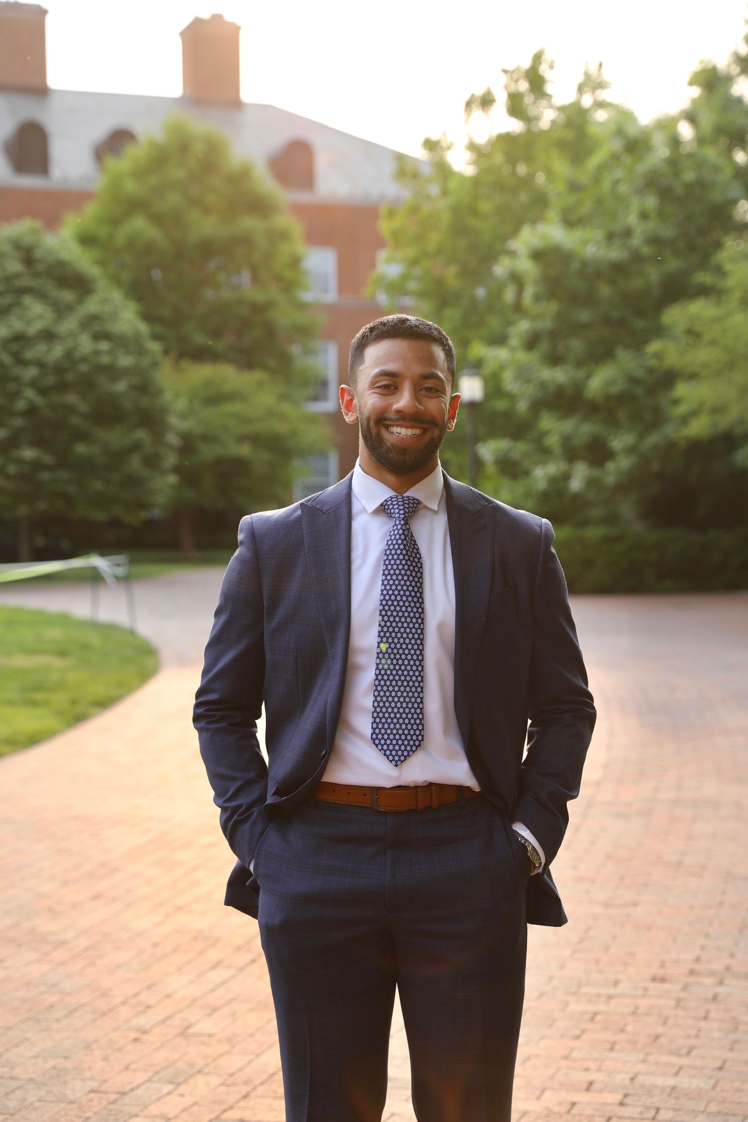Professional wearing a navy suit and polka dot tie stands confidently on brick pavement with buildings in background.