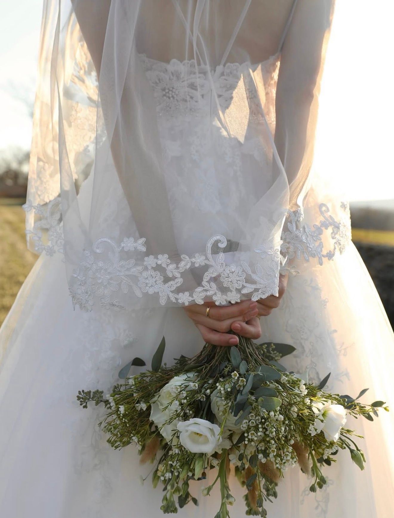 Close-up detail of delicate white lace wedding dress with floral appliques and greenery bouquet accents.