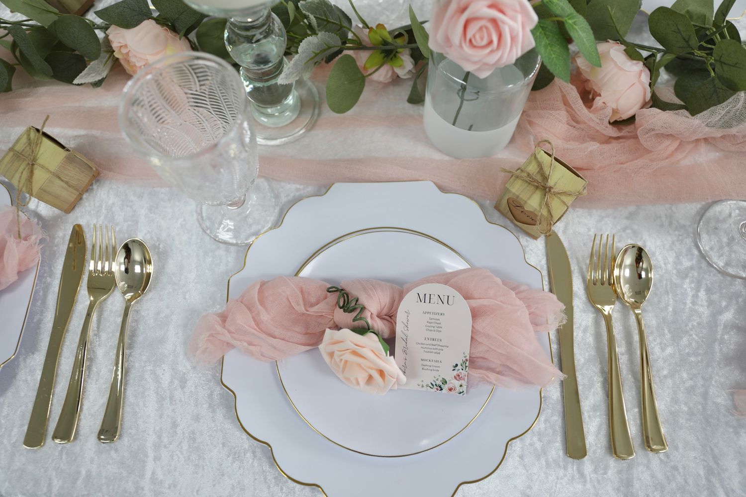 Elegant table setting with pink roses, eucalyptus, gold cutlery, and glass jars creating a romantic wedding reception ambiance.