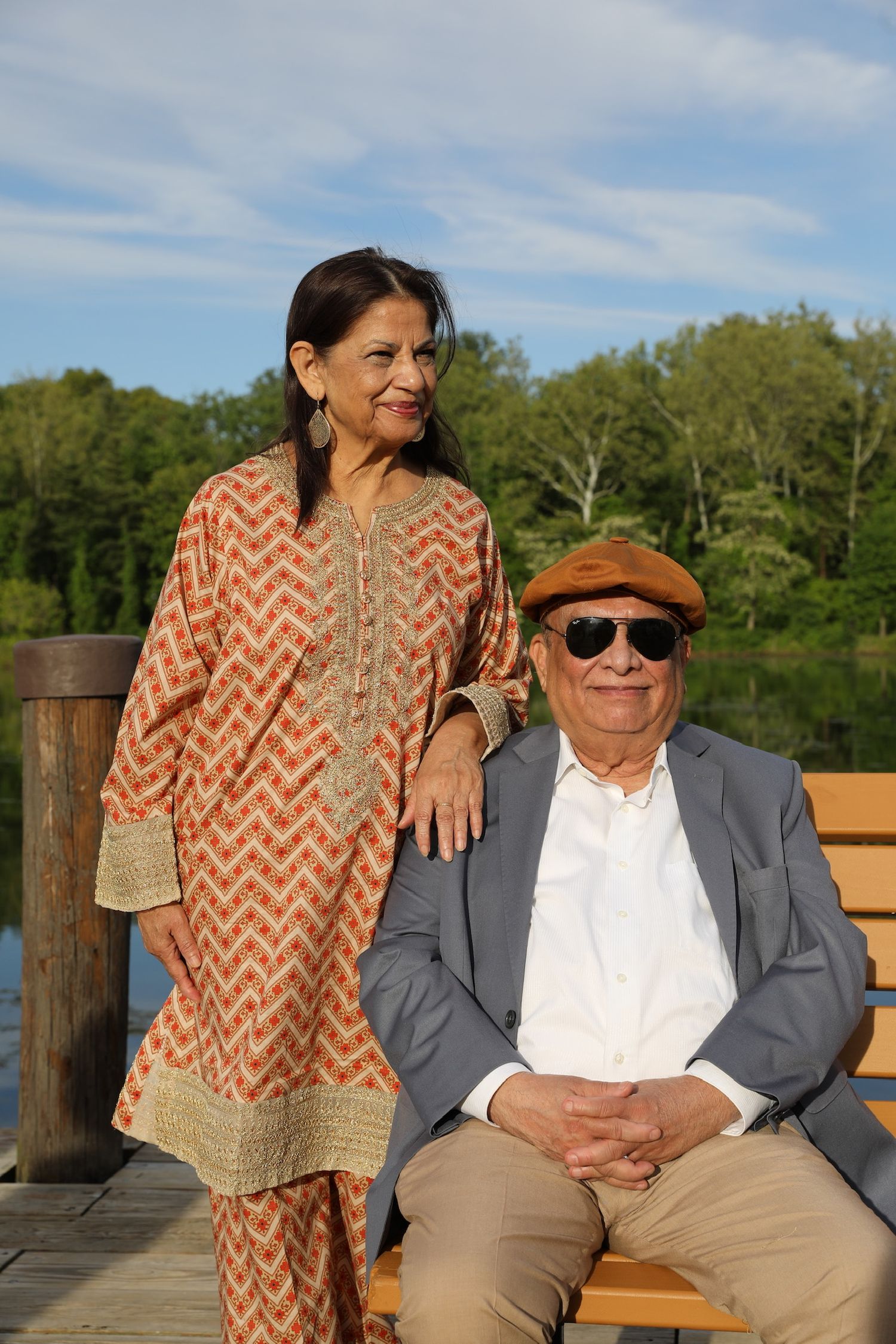 Two people sit together on a wooden bench by a lake, wearing casual attire on a sunny day.