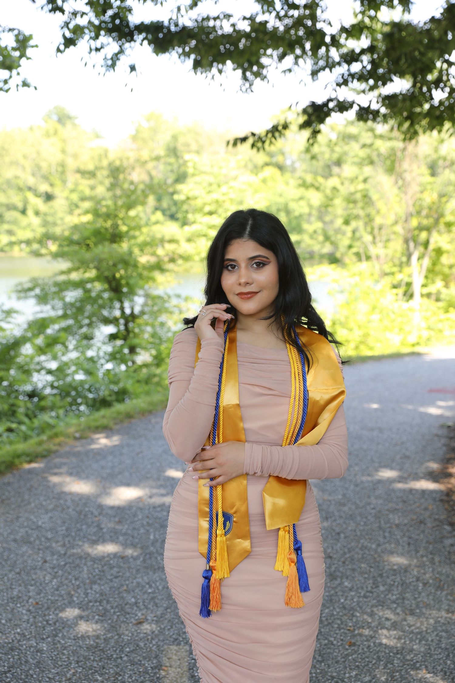A person poses outdoors wearing a beige dress and yellow graduation stole with blue tassels in a natural setting.