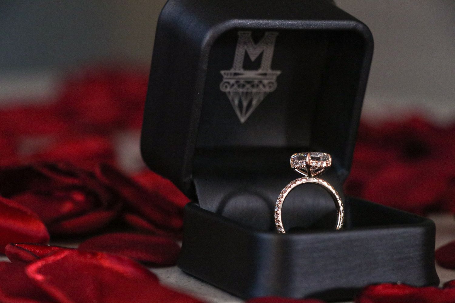 A sparkling diamond engagement ring sits in a black box surrounded by red rose petals.