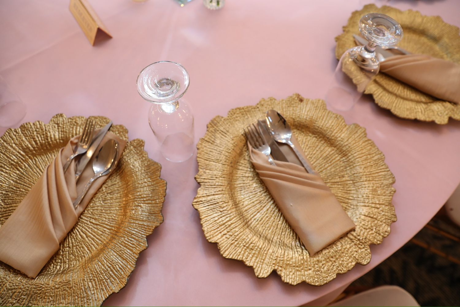 Gold scalloped charger plates with tan napkins arranged on a pink tablecloth for an elegant table setting.