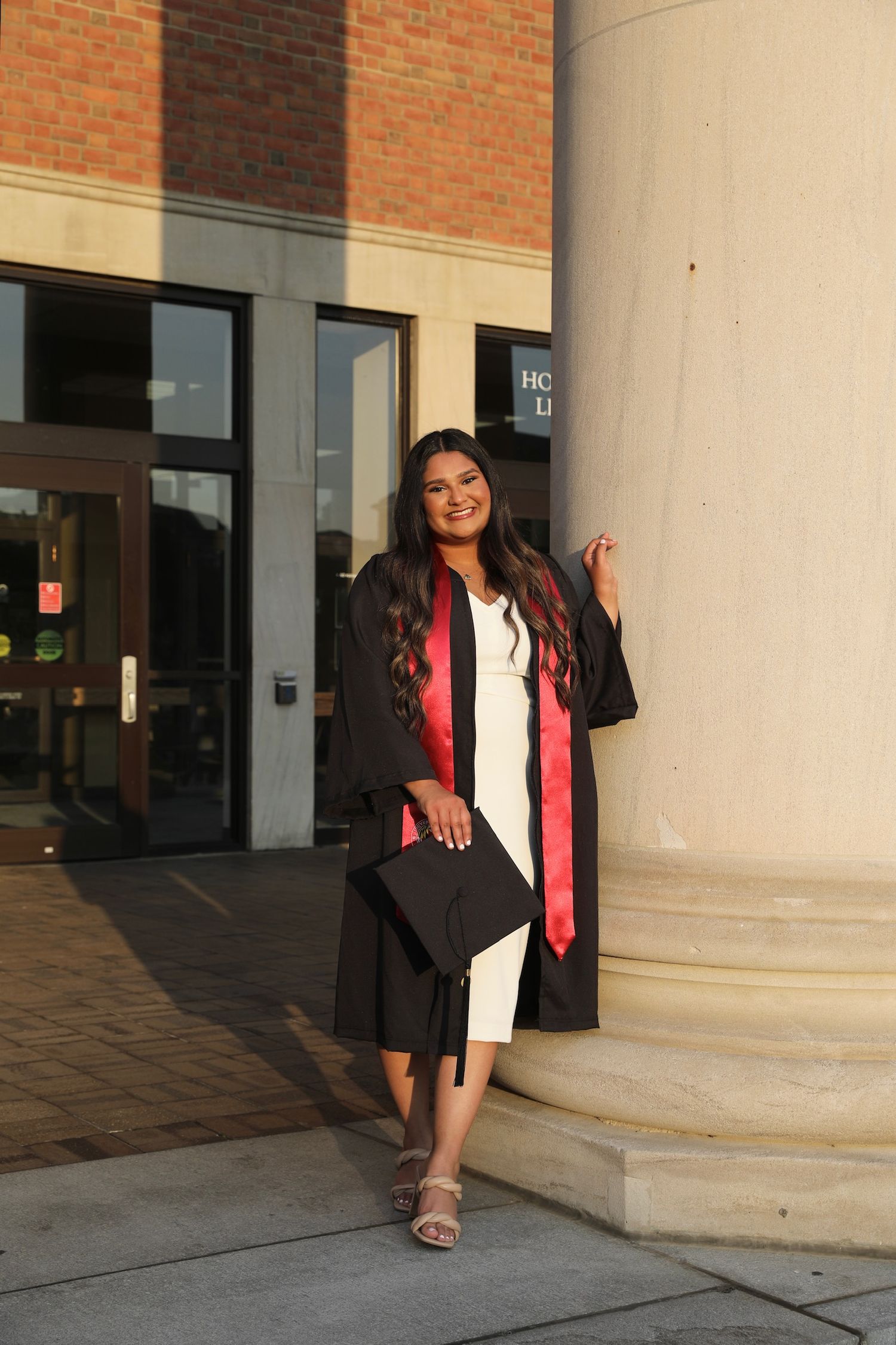 A graduate in black cap and gown with pink stole poses next to a white column in a series of photos.