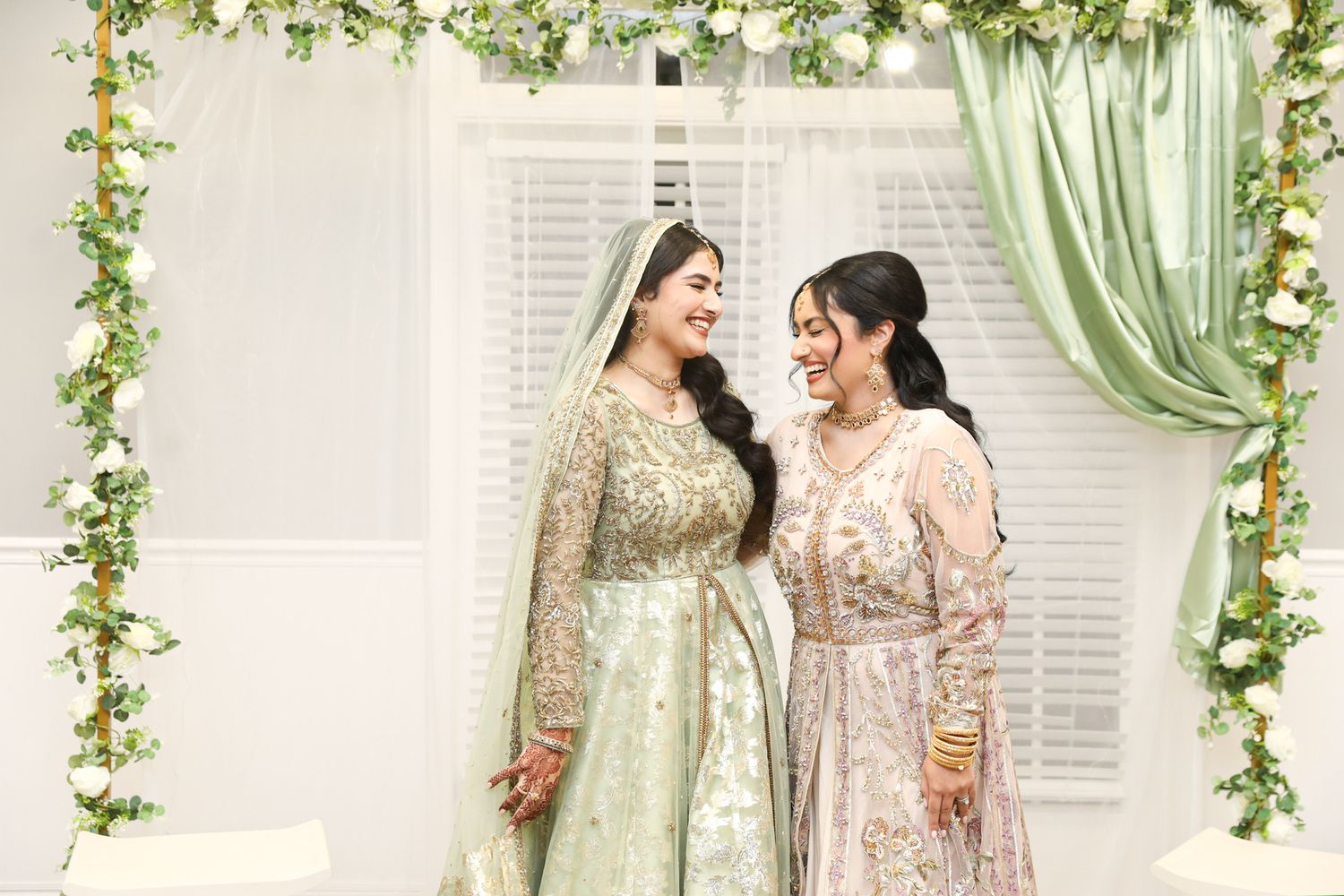 Wedding ceremony backdrop decorated with elegant white floral arrangements and soft green drapery against a window.
