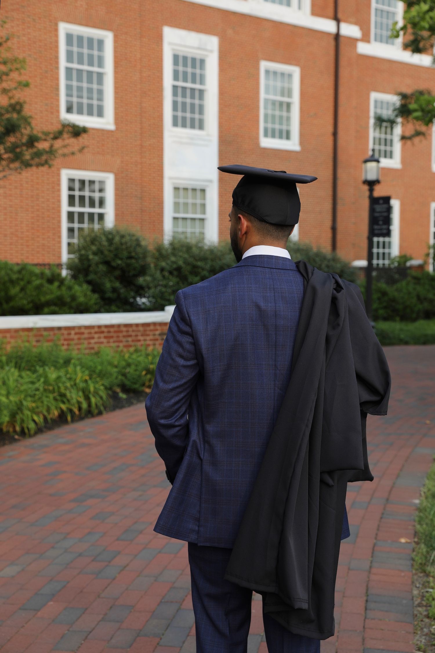 A graduate in cap and gown walks along a brick path in front of a red brick university building.